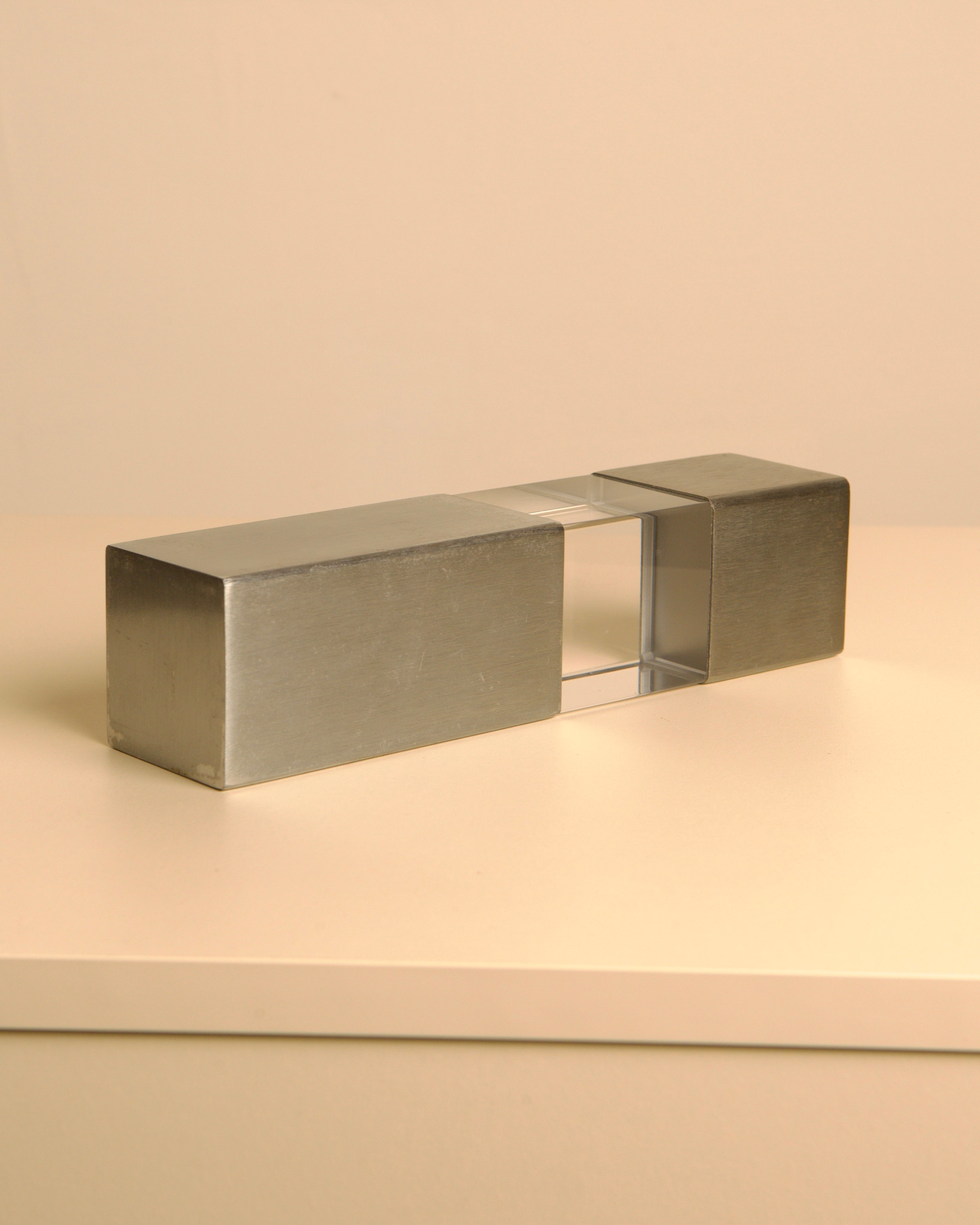 The Treaptyque Italian Sculpture in Metal and Plexiglass 70's features brushed metal ends and a transparent plexiglass center, displayed on a light surface with a beige background, reflecting the sleek elegance of 1970s Italian design.