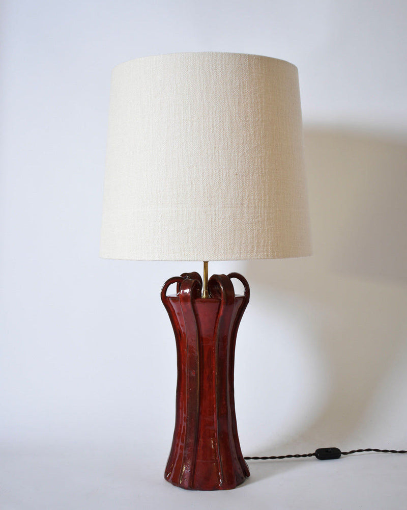 The "Kasaï" Henry Red Ceramic Lamp by Barracuda Interiors features a tall, glossy red base with curved accents and an off-white fabric shade. Shown plugged in against a plain white background.