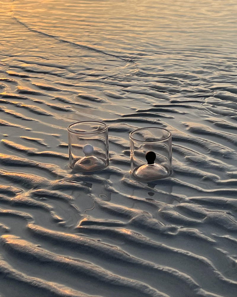 Two clear glass shot glasses with black and white spheres on textured sand beach