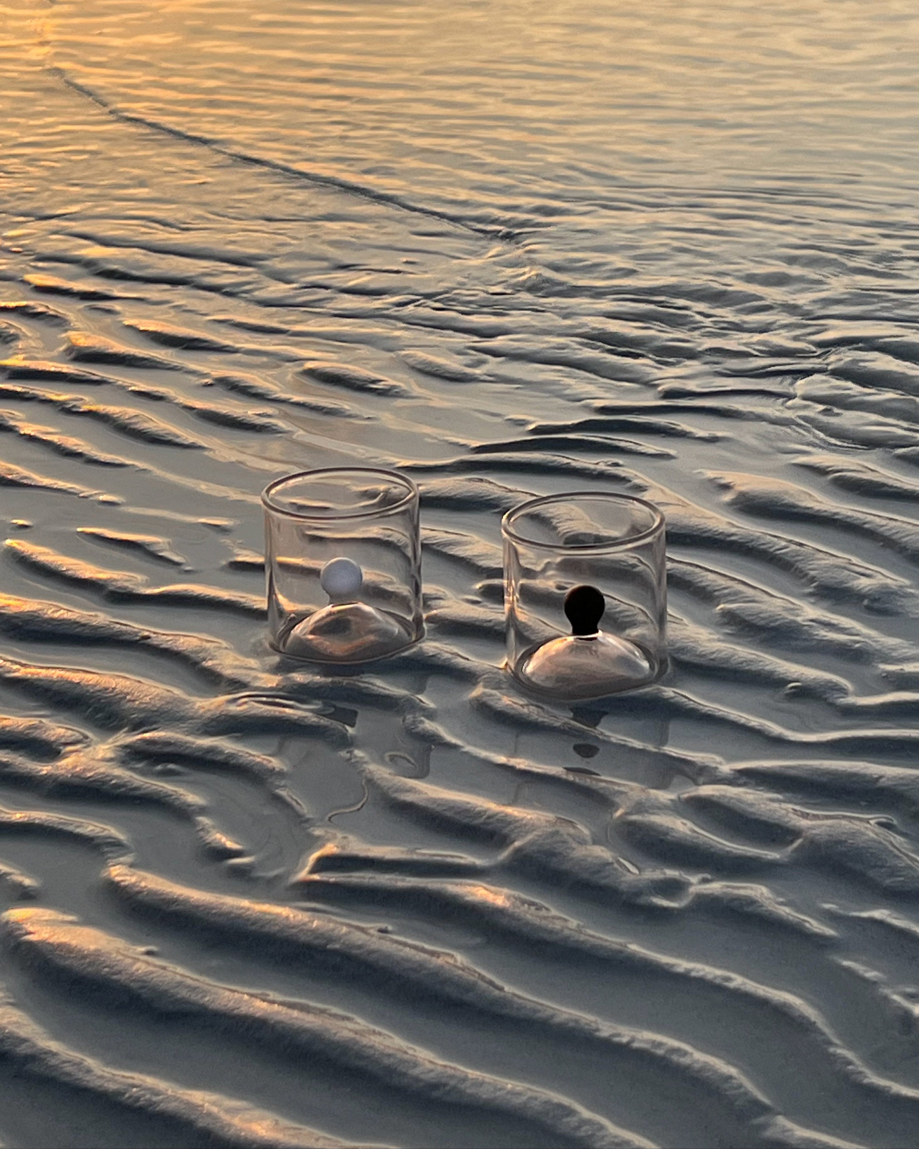 Two clear glass shot glasses with black and white spheres on textured sand beach