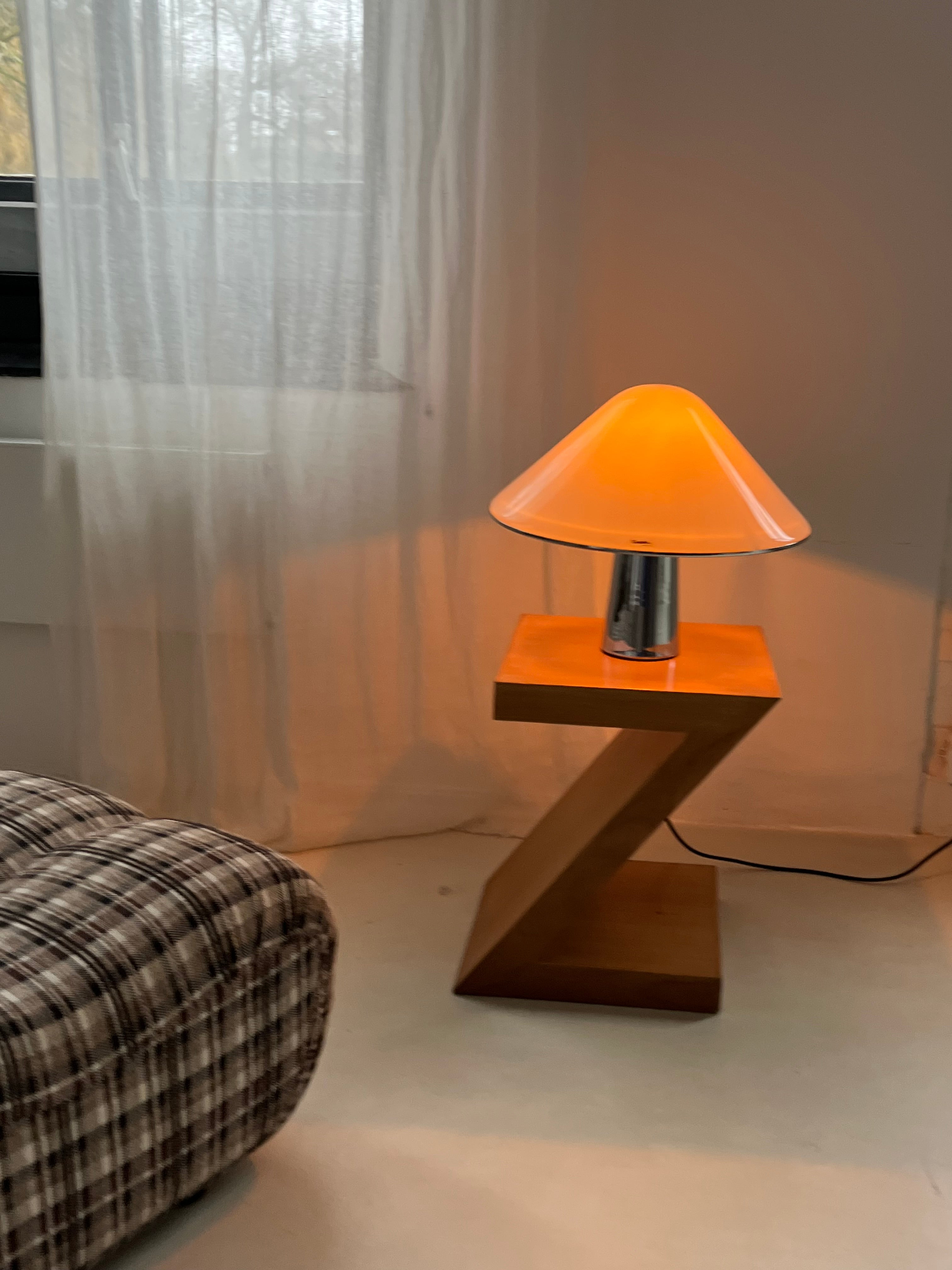 A modern room features a wooden Z-shaped side table with the Harvey Guzzini Elpis Table Lamp by septembre studios, its orange shade beside a plaid sofa and a window dressed with sheer white curtains.