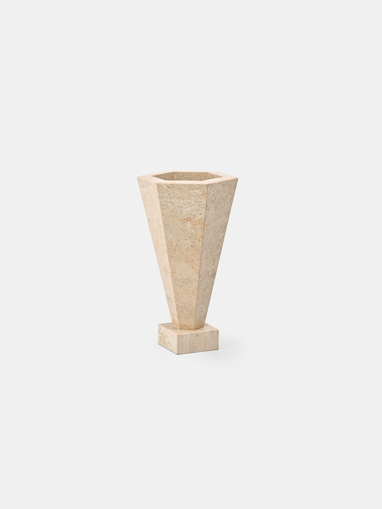 The Vase 01 Travertine by Studio Vraco, featuring a tall hexagonal design and narrow base in beige travertine, stands on a small square pedestal against a plain white background.