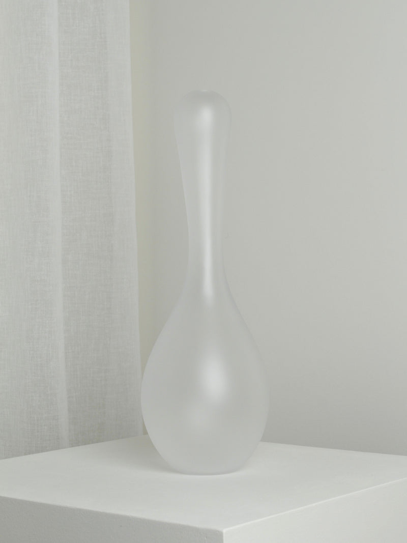 The XLarge Vase in Ghost by Verre d'Onge, featuring frosted glass with a narrow neck and bulbous base, rests on a white pedestal set against a light gray wall and sheer curtain.