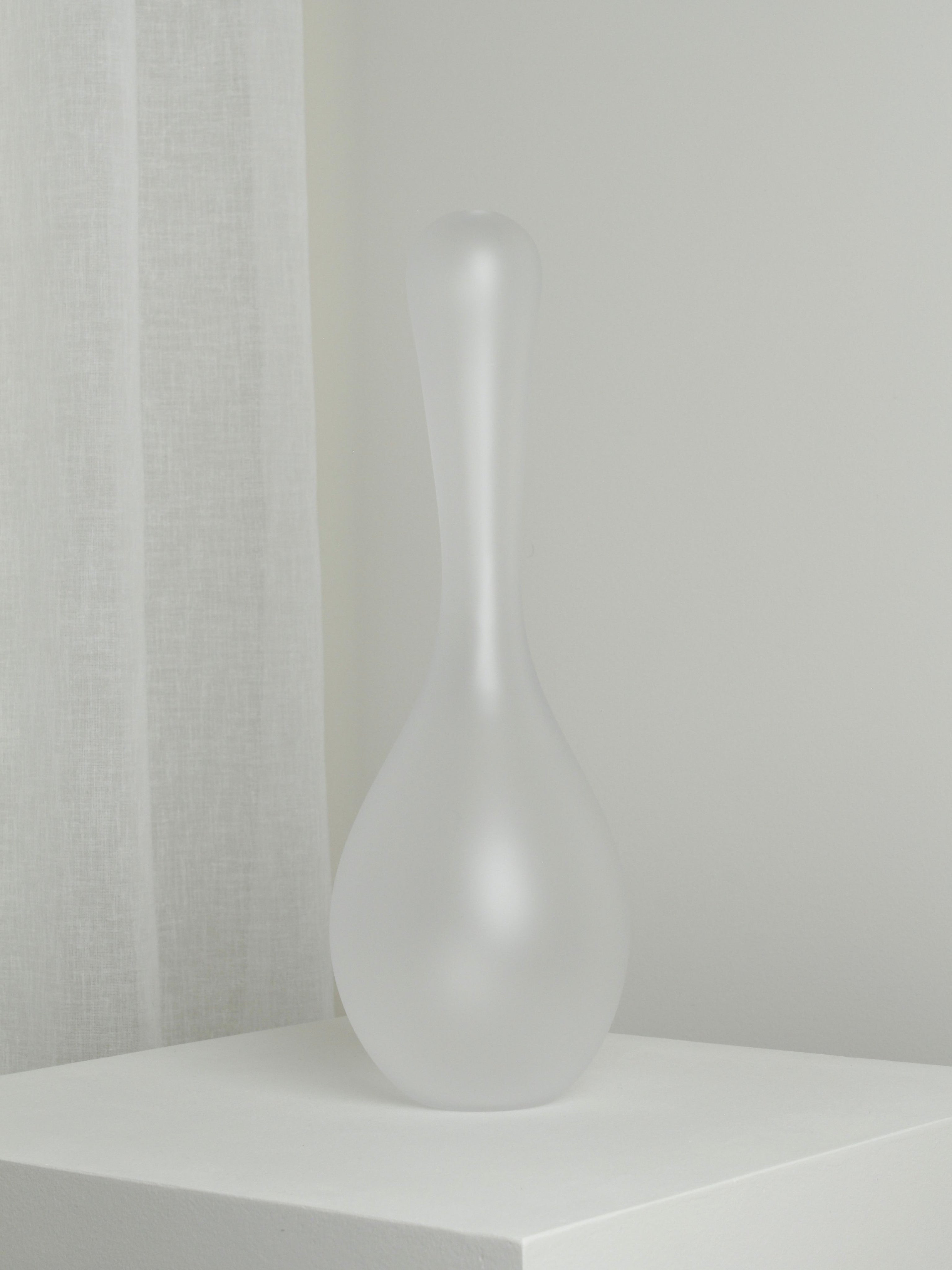 The XLarge Vase in Ghost by Verre d'Onge, featuring frosted glass with a narrow neck and bulbous base, rests on a white pedestal set against a light gray wall and sheer curtain.