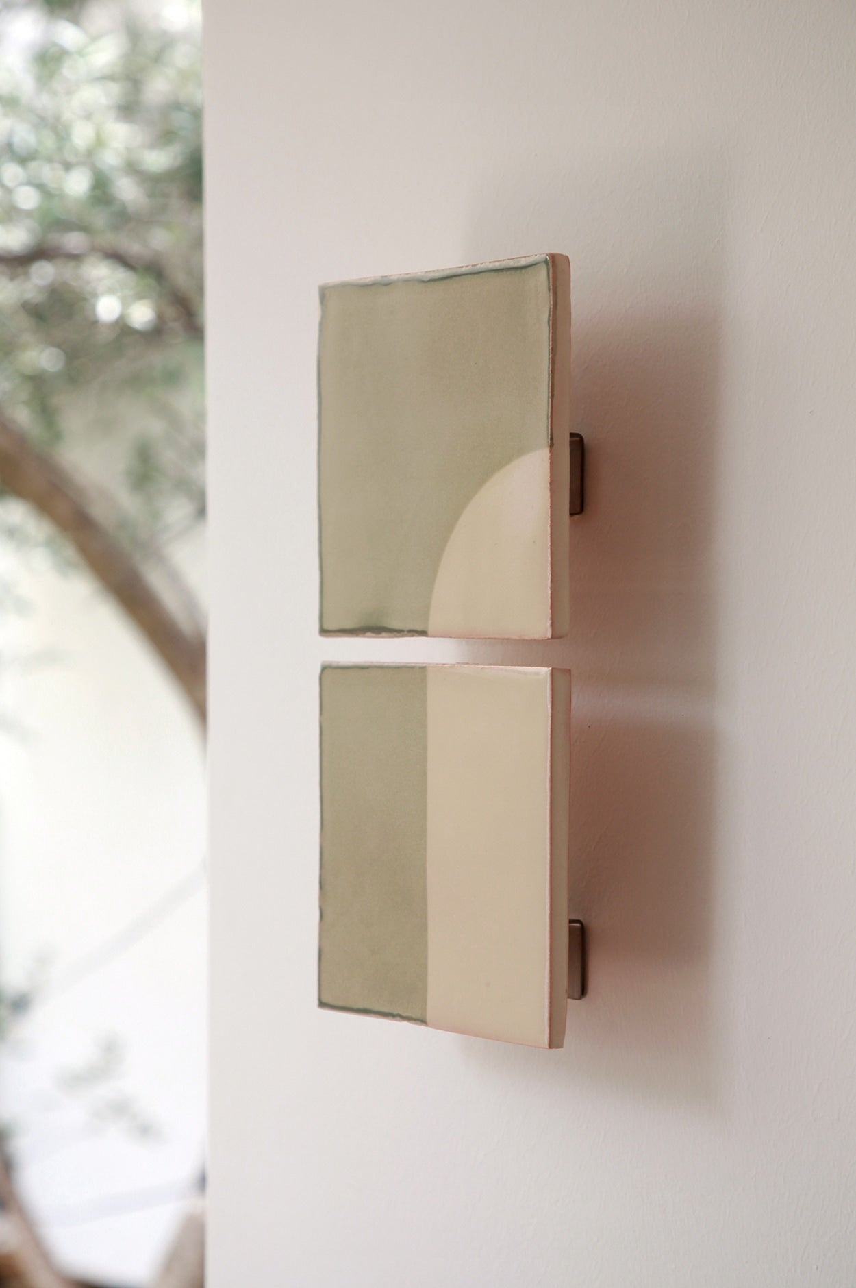 Two minimalist Tiles Door V appliques murales céramique by Violaine d'Harcourt, featuring geometric designs in muted green and cream, are mounted on a white wall. Soft light and a blurred leafy backdrop highlight this artisan décor.