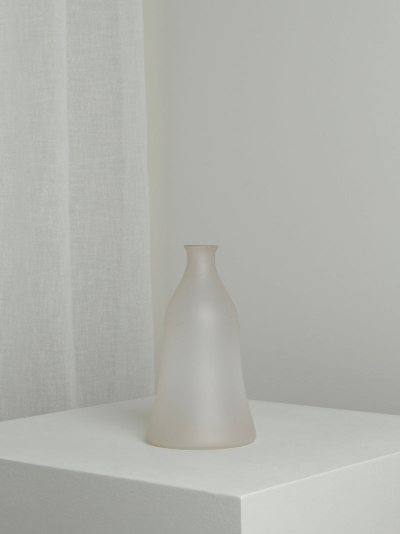 The Medium Vase in Moon by Verre d'Onge, featuring frosted glass and a narrow neck, is displayed on a white pedestal against a light gray wall and sheer curtain in a minimalistic, softly lit setting.