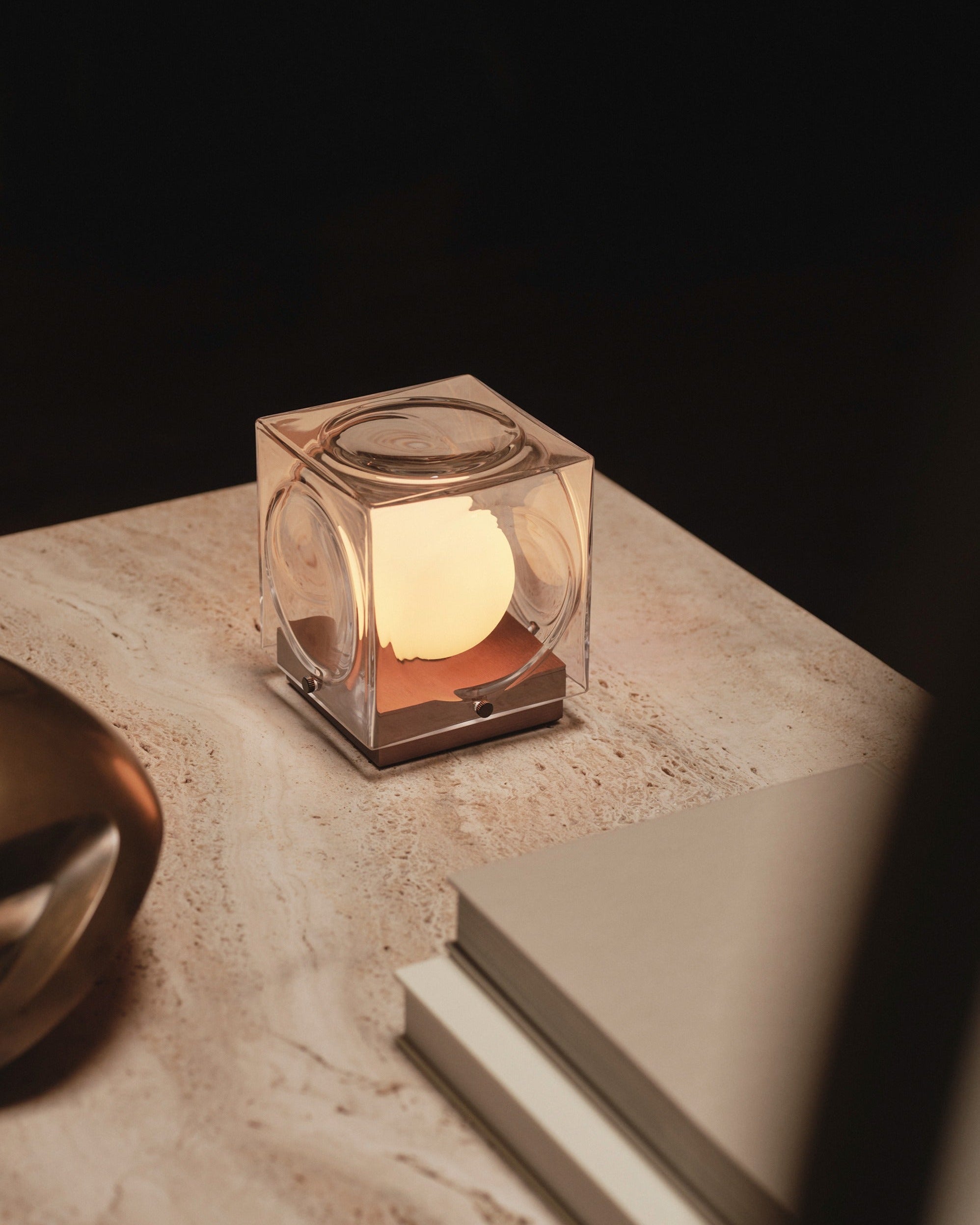 The Lee Broom Chant Portable Table Lamp, a compact cube-shaped lamp with an inner glow, is displayed on a marble surface beside a closed book and metallic decor in soft lighting.