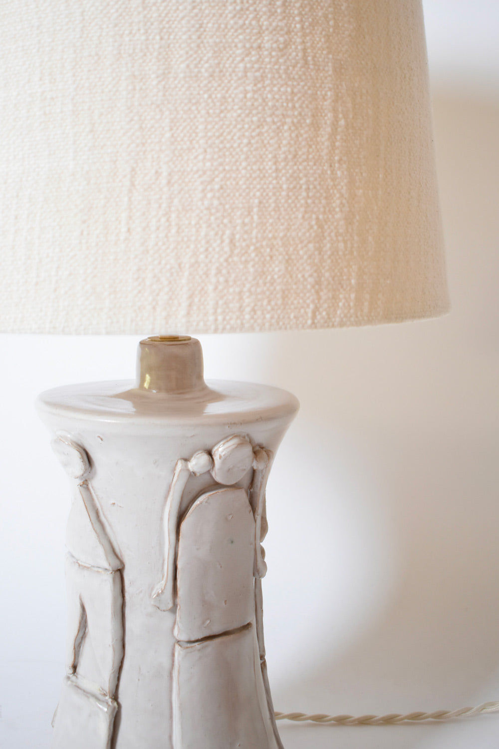 The "Kasaï" Avignon Ceramic Table Lamp by Barracuda Interiors features a textured off-white ceramic base with two abstract human figures and a cream-colored, textured fabric lampshade—perfect for customizable interior design.