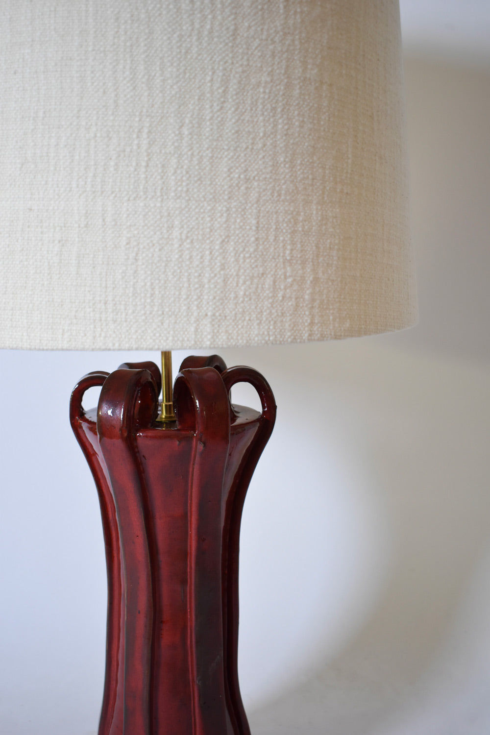 A close-up of the "Kasaï" Henry Red Ceramic Lamp by Barracuda Interiors, featuring Albarelo-inspired curves, a brass fixture, and a large textured off-white shade, set against a plain background.