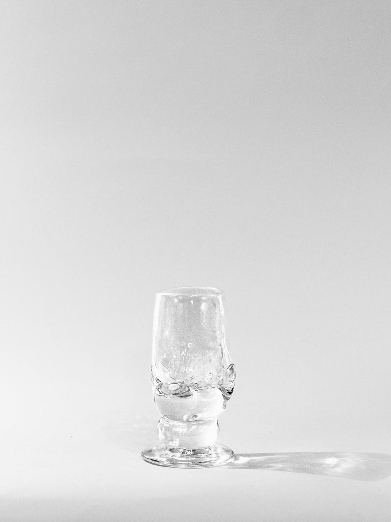An empty Szkło Studio Organic Footed Table Glass stands upright on a white surface, casting a faint shadow to the right against a plain, light background.