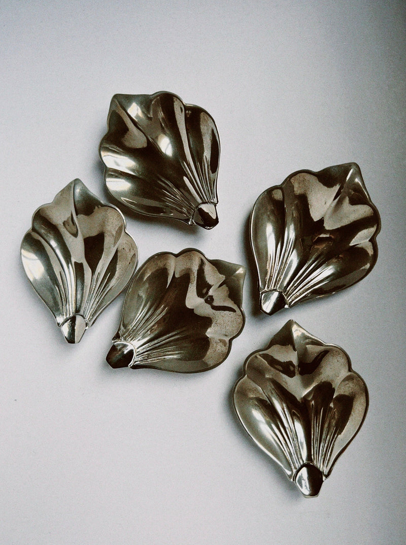 The Manushi Silver Table Decor Set features five shiny, scallop-shaped baking molds with seashell-like ridges, arranged in a loose circle to add a charming touch to any setting.