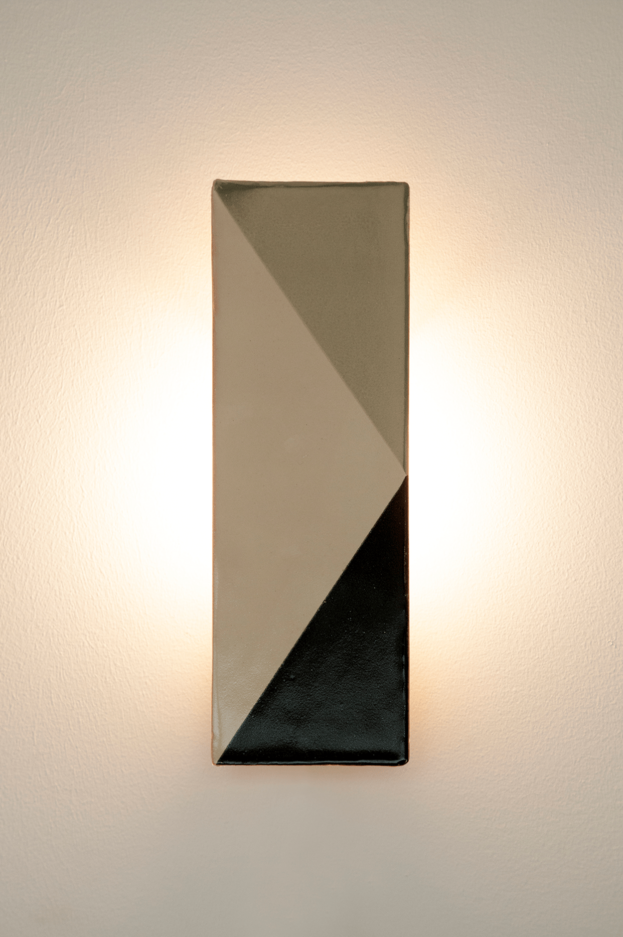 The Tile VN rectangular wall sconce by Violaine d'Harcourt features a geometric black, gray, and beige pattern and emits soft indirect light, creating a glowing effect on the white wall. This handmade ceramic fixture is crafted in Portugal.