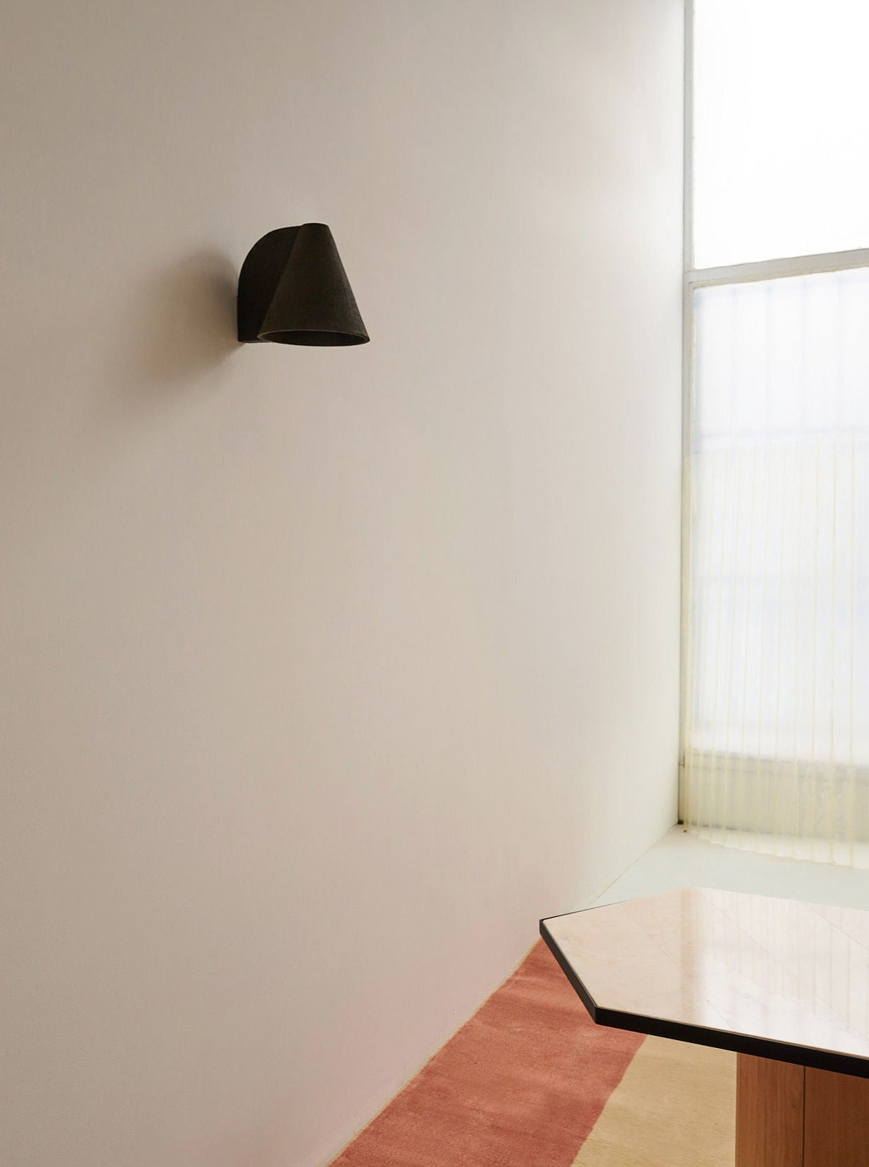 Minimalist interior with a white wall, Mariza Galani's Esoteric Wall Lamp in black adding modern lighting, a hexagonal table with a light top, and a geometric rug beside a tall window dressed with sheer curtains.
