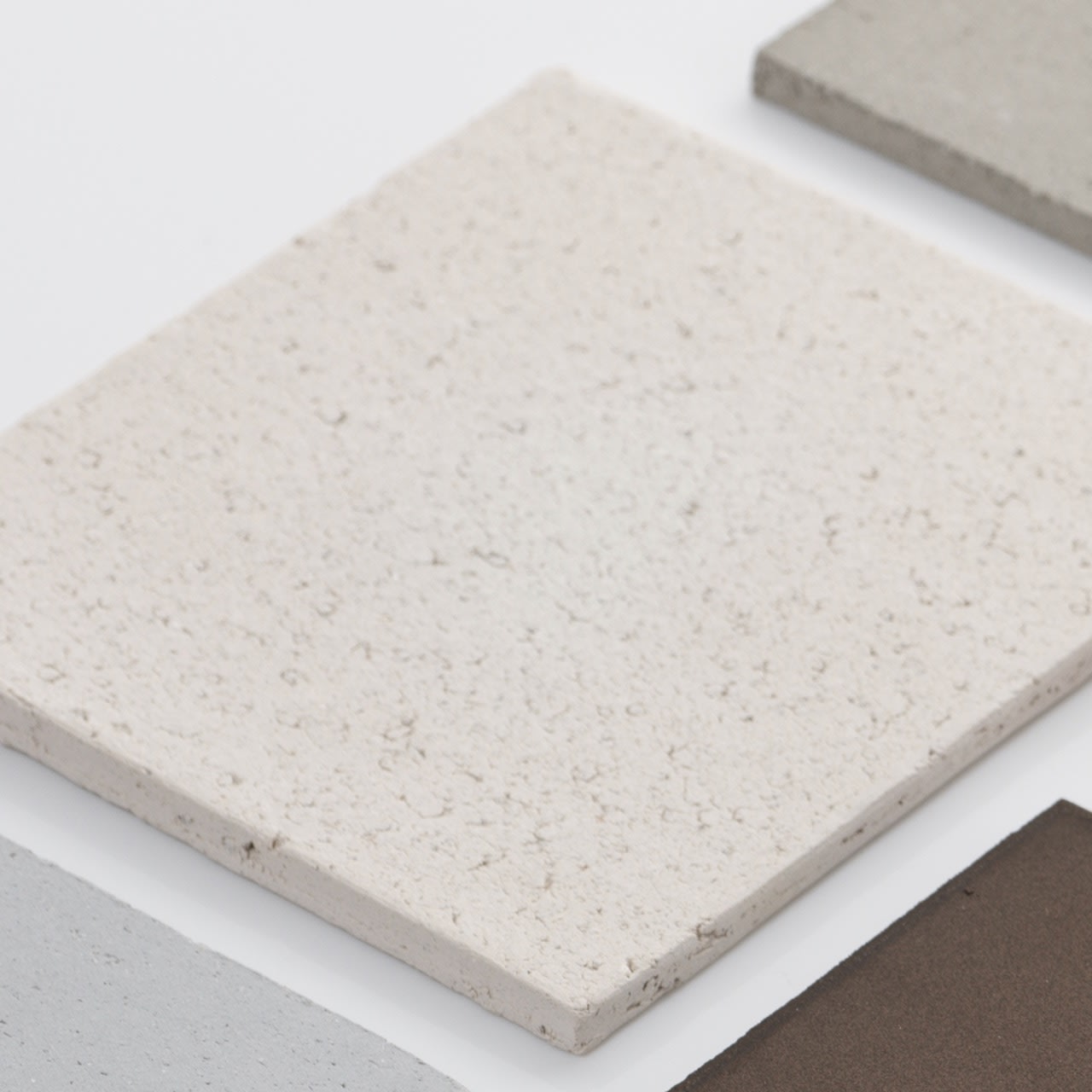 A close-up of four square tiles in off-white, light gray, beige, and dark brown rests on a white surface next to the 'Too 60' Ceramic Table Lamp by Katia Langeard.