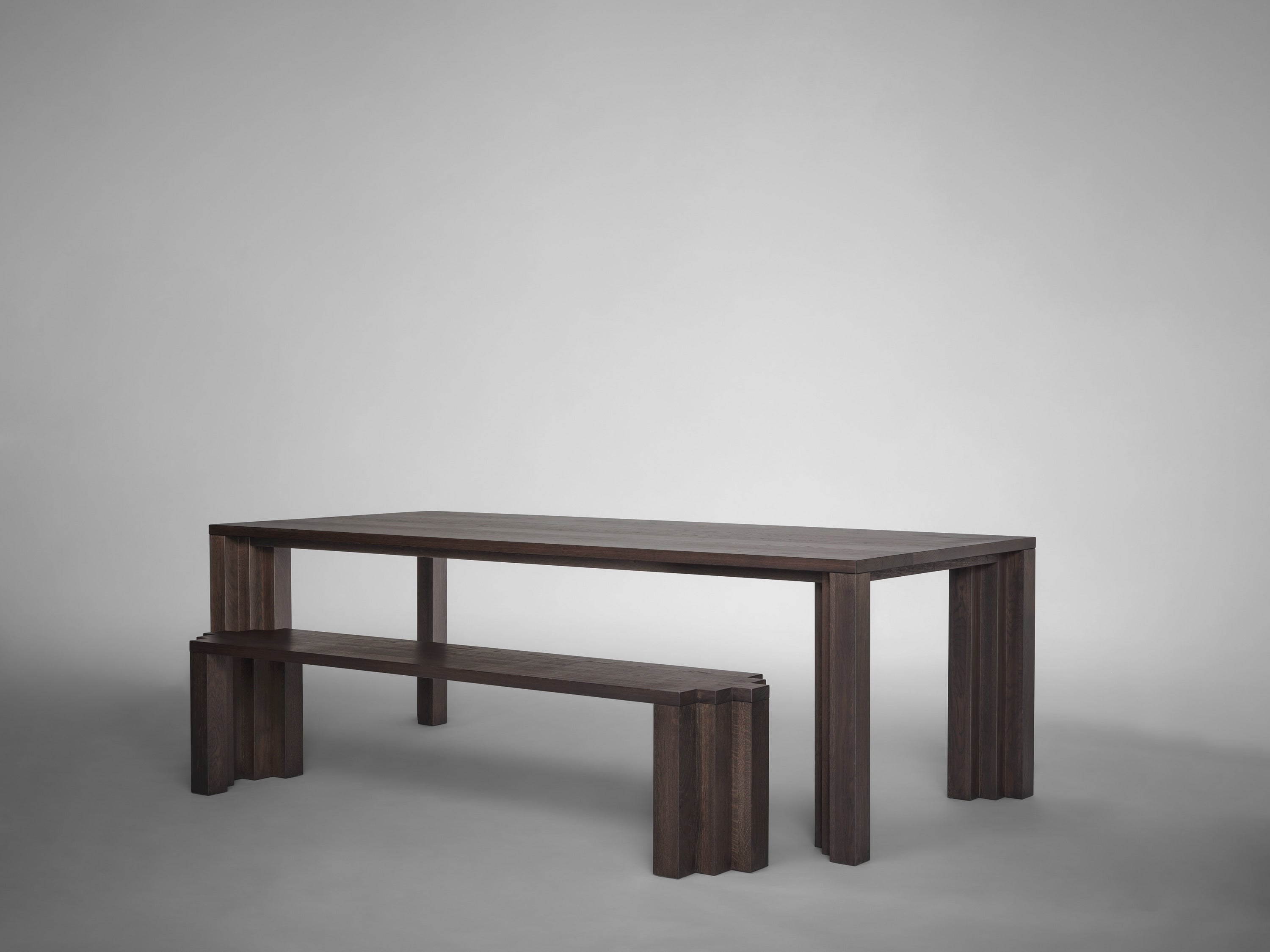 A dark wooden Mokko Cadence Bench fits under a matching table, both with thick fluted legs. Set against a plain gray backdrop, this minimalist furniture showcases customizable solid European oak craftsmanship.
