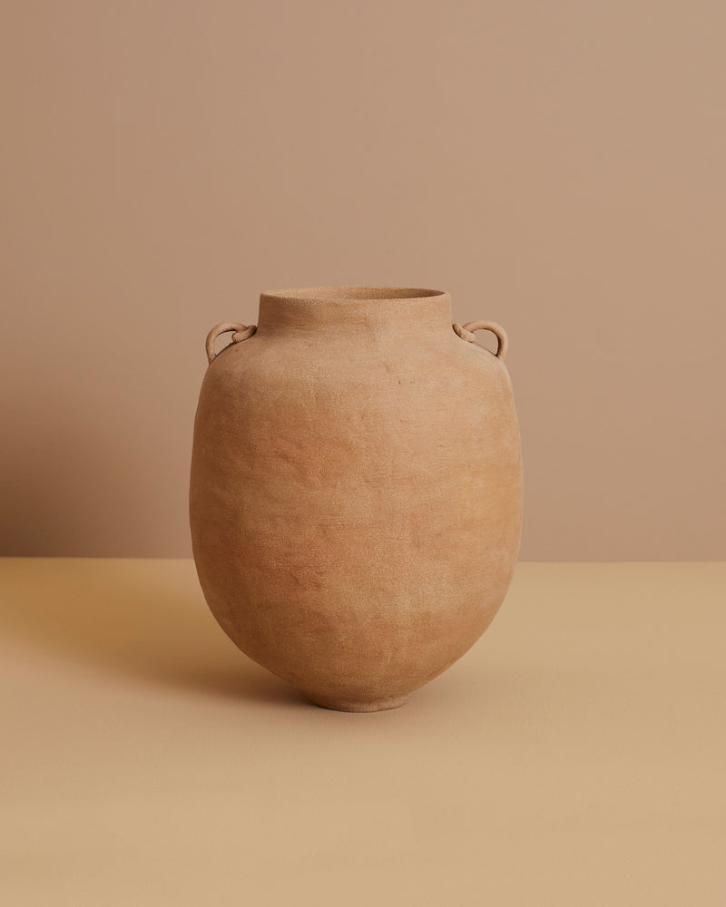 A large, round Column Vase by marta bonilla in light brown terracotta stoneware with two small handles elegantly rests on a beige surface against a matching background.