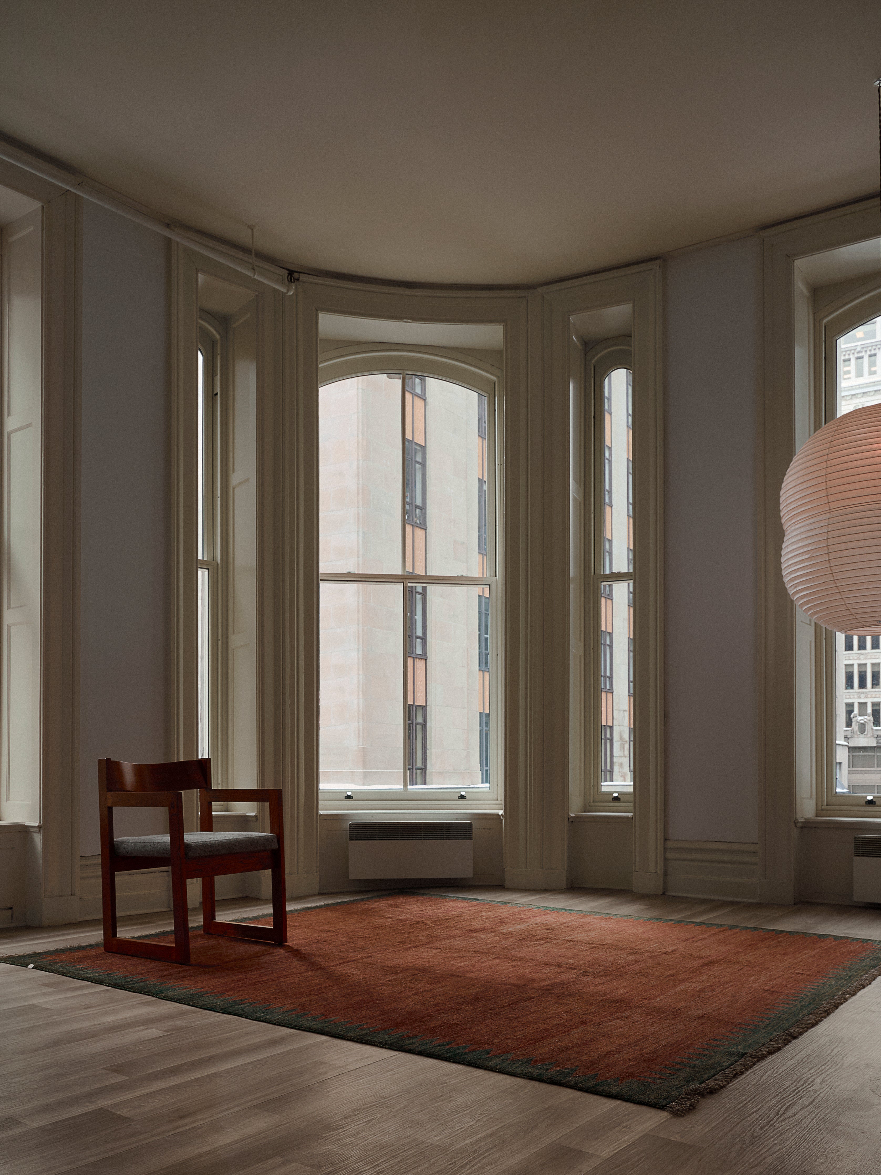 The minimalist room features tall windows allowing soft light to flood in. A wooden chair sits on a catalog:three Ziegler Rug in Carob (244 cm x 274 cm). Overhead, a round pendant lamp illuminates, while the view reveals a nearby building, crafting a simple yet modern atmosphere.