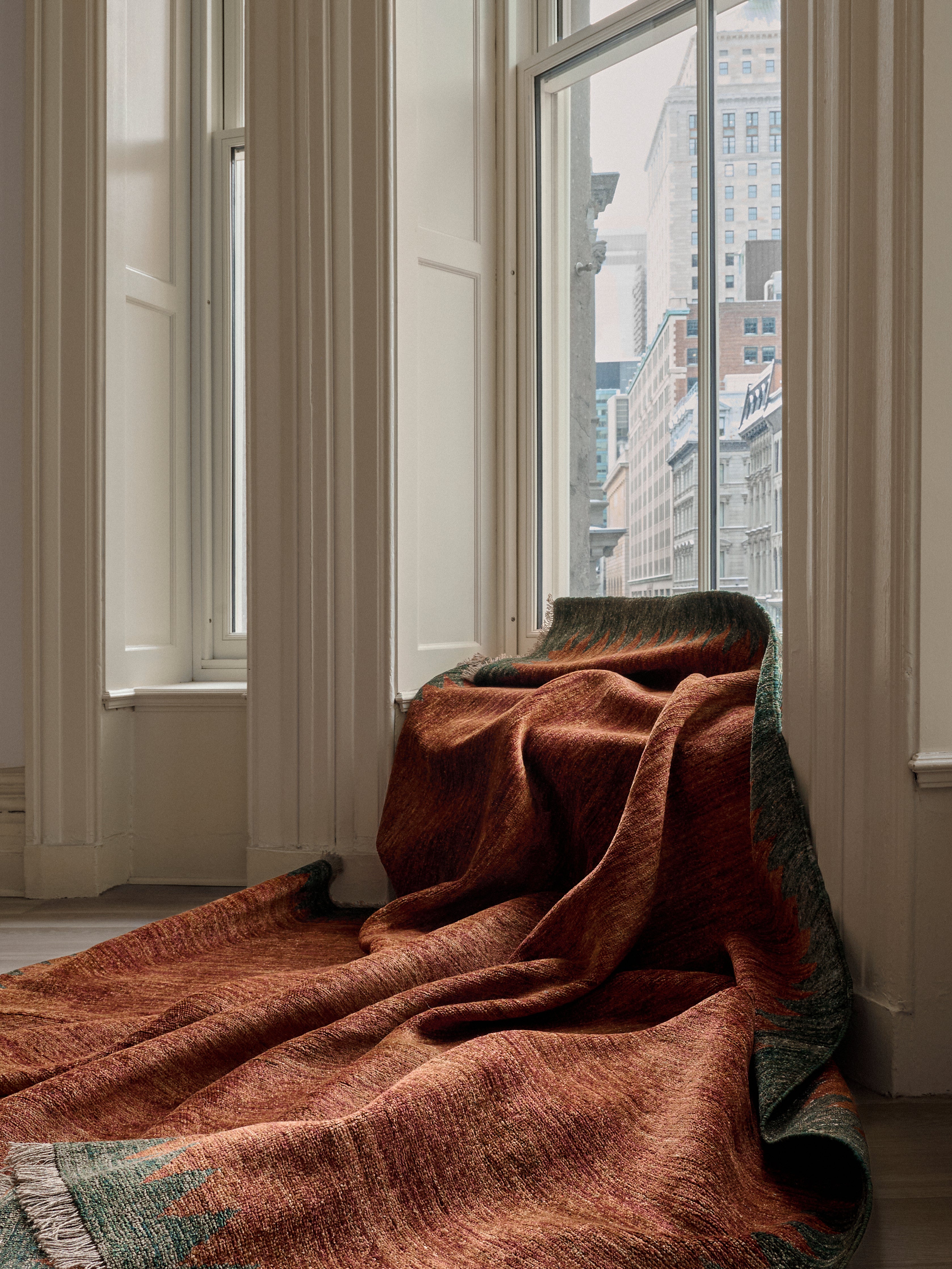 A rust woven blanket drapes over the ledge of a white-paneled bay window, complementing a hand-knotted Ziegler Rug in Carob by catalog:three. Tall buildings outside contrast with the cozy setting, while the blanket adds warmth and texture to the serene space.