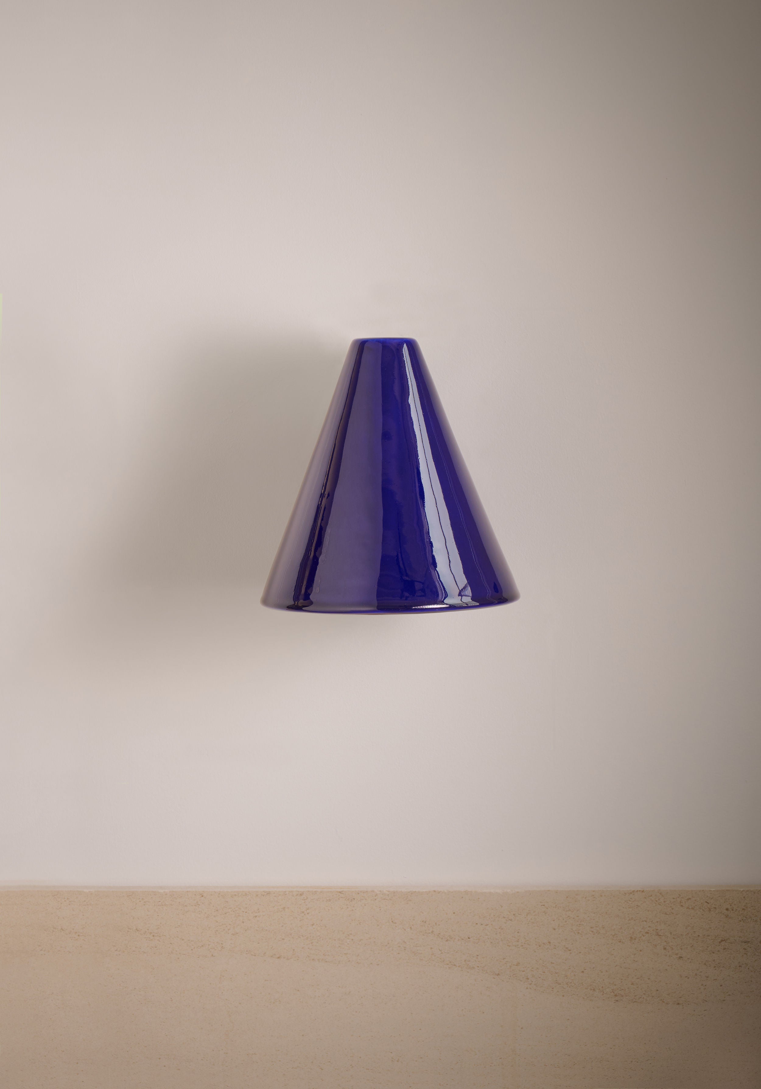 The 'Esoteric' Blue Wall Light by Mariza Galani, a glossy handcrafted ceramic sconce in midnight blue, is mounted on a plain light beige wall above pale wood, creating a minimalist and modern look.