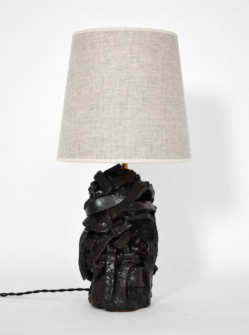 The Small "Angkor" Black Glazed Terracotta Lamp by Barracuda Interiors features a sculptural, textured base and a beige fabric shade, displayed against a plain white background.
