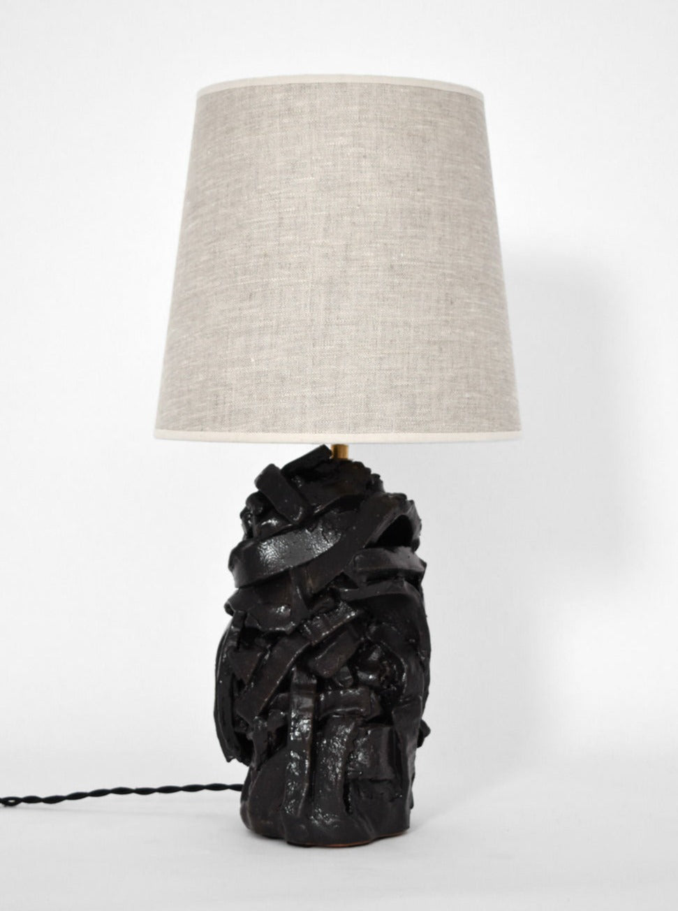 The Small "Angkor" Black Glazed Terracotta Lamp by Barracuda Interiors features a sculptural, textured base and a beige fabric shade, displayed against a plain white background.