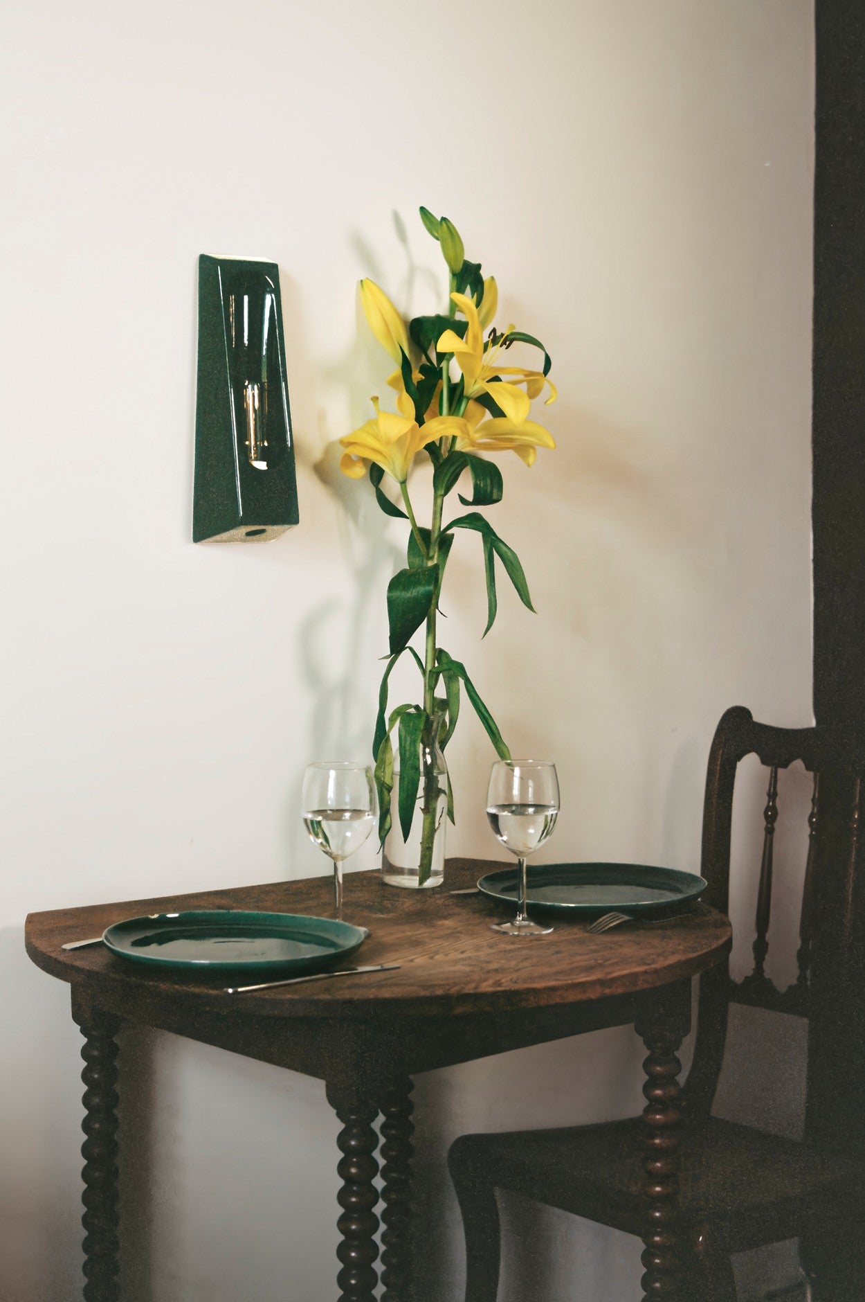 The Alcove Verde dining set by Violaine d'Harcourt seats two and includes green plates, cutlery, wine glasses, and a vase of yellow lilies, all softly lit by elegant interior lighting from nearby alcoves.