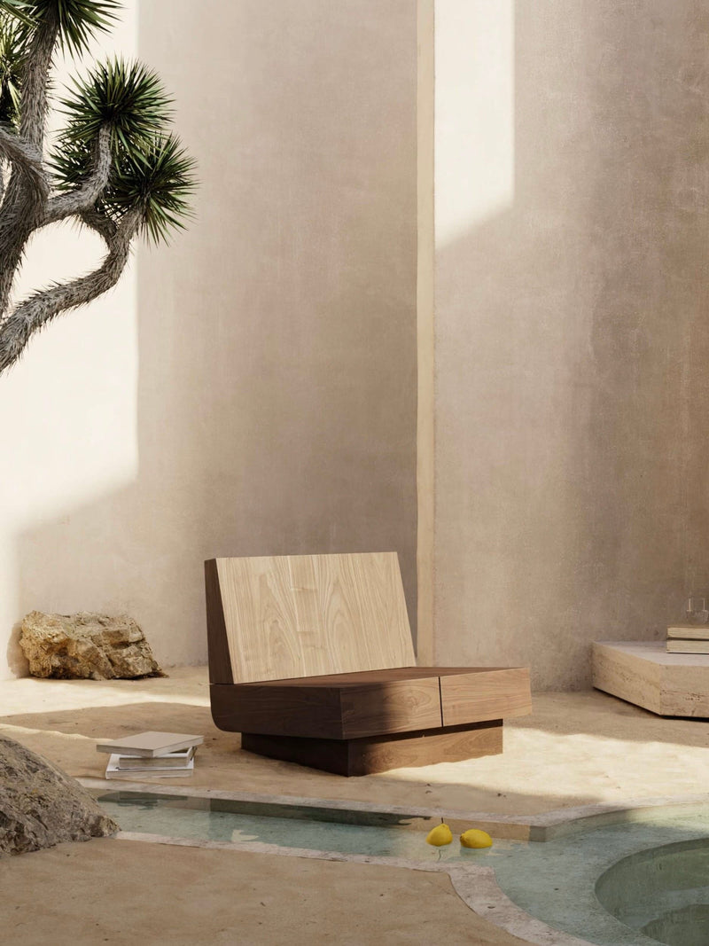 A minimalist M_007 Lounge Chair / Walnut by Monolith sits beside a small pool in a sunlit courtyard with beige stone walls, a rock, spiky-leaved tree, and two lemons on the ground, creating a calm and serene scene.
