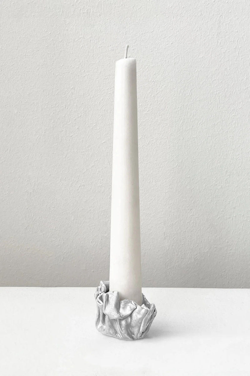 A tall white taper candle stands in the Completedworks L42 - Candle Holder in Silver, its metallic crumpled design shown against a plain white background.