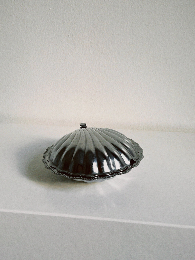 The Manushi Shell Shaped Butter Dish, featuring a silver finish, scalloped edge, and small handle, sits on a white surface against a plain light-colored wall.
