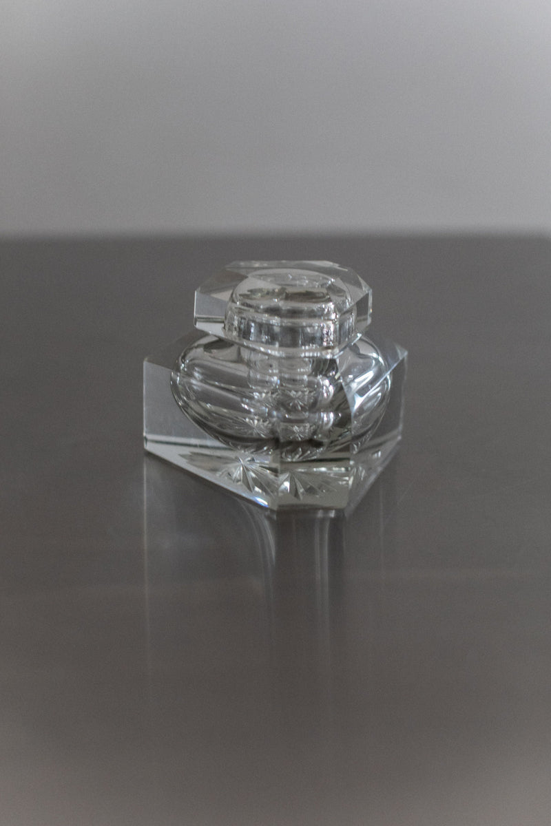 A clear, faceted Glass Perfume Bottle by Out For Lunch, featuring exquisite craftsmanship and a matching lid, sits on a reflective gray surface, casting subtle shadows and reflections.