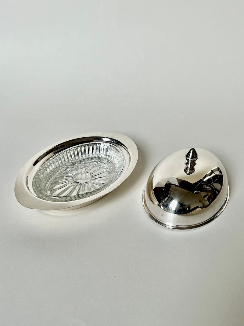 The Manushi Silver Butter Dish features an oval shape with a decorative glass insert and a matching silver lid topped with a pointed knob, all presented against a plain white background.