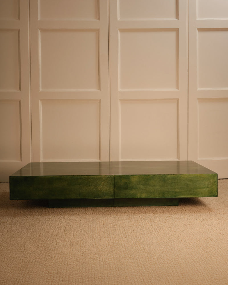 A Green parchment table by Galerie Vauvart, with a glossy rectangular surface, sits on a beige carpet before a cream-paneled wall in a softly lit, minimal space that echoes Aldo Tura’s signature style.