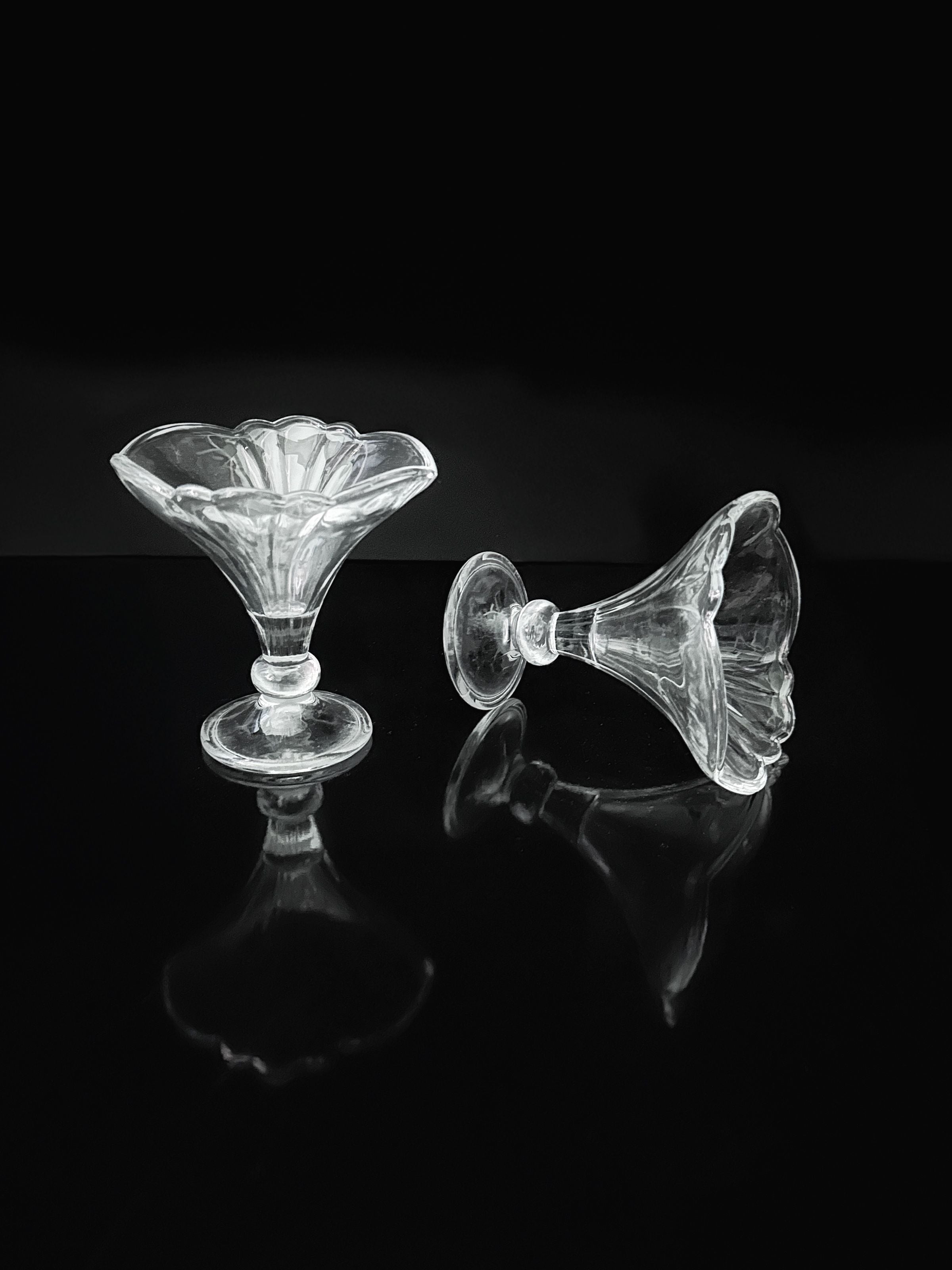Four Fleur-De-Lis stylised dessert cups by Dodo Vintage are displayed—one upright, another on its side—on a glossy black surface, their reflections evoking Art Deco Revival sophistication.