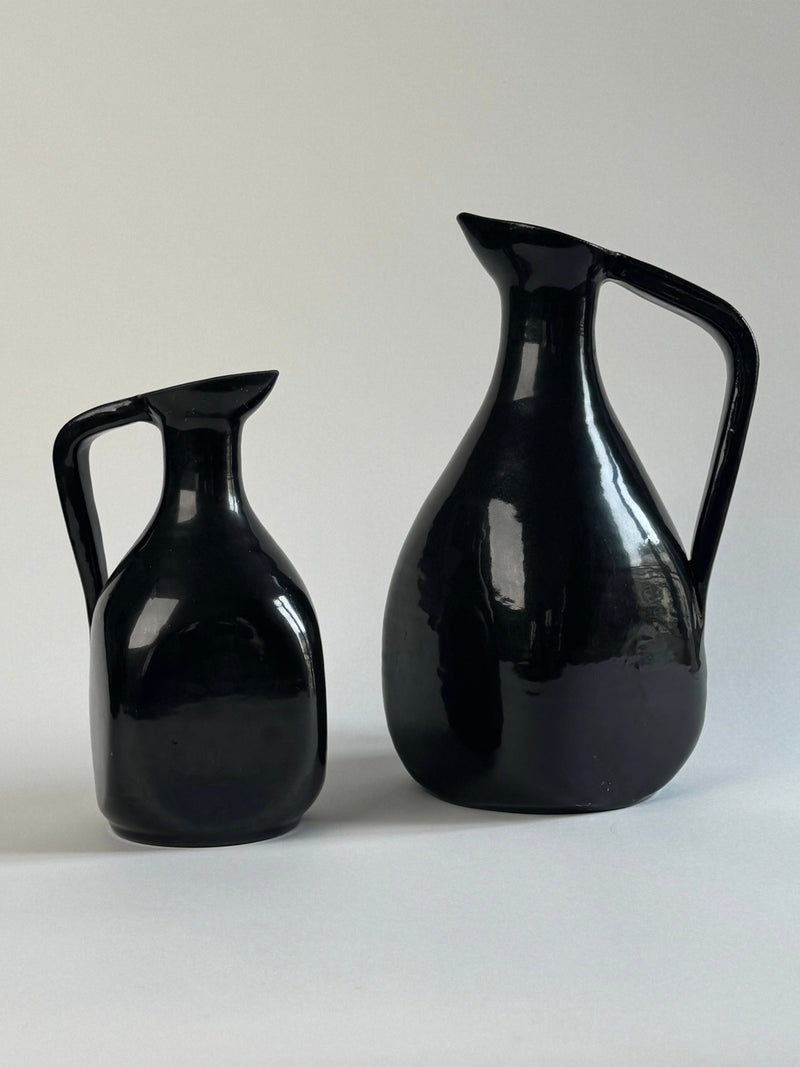 A Pair of Accolay Ceramic Carafes by Figuresdesign, featuring glossy black curved bodies and handles—one large, one small—are displayed side by side against a neutral light gray background.