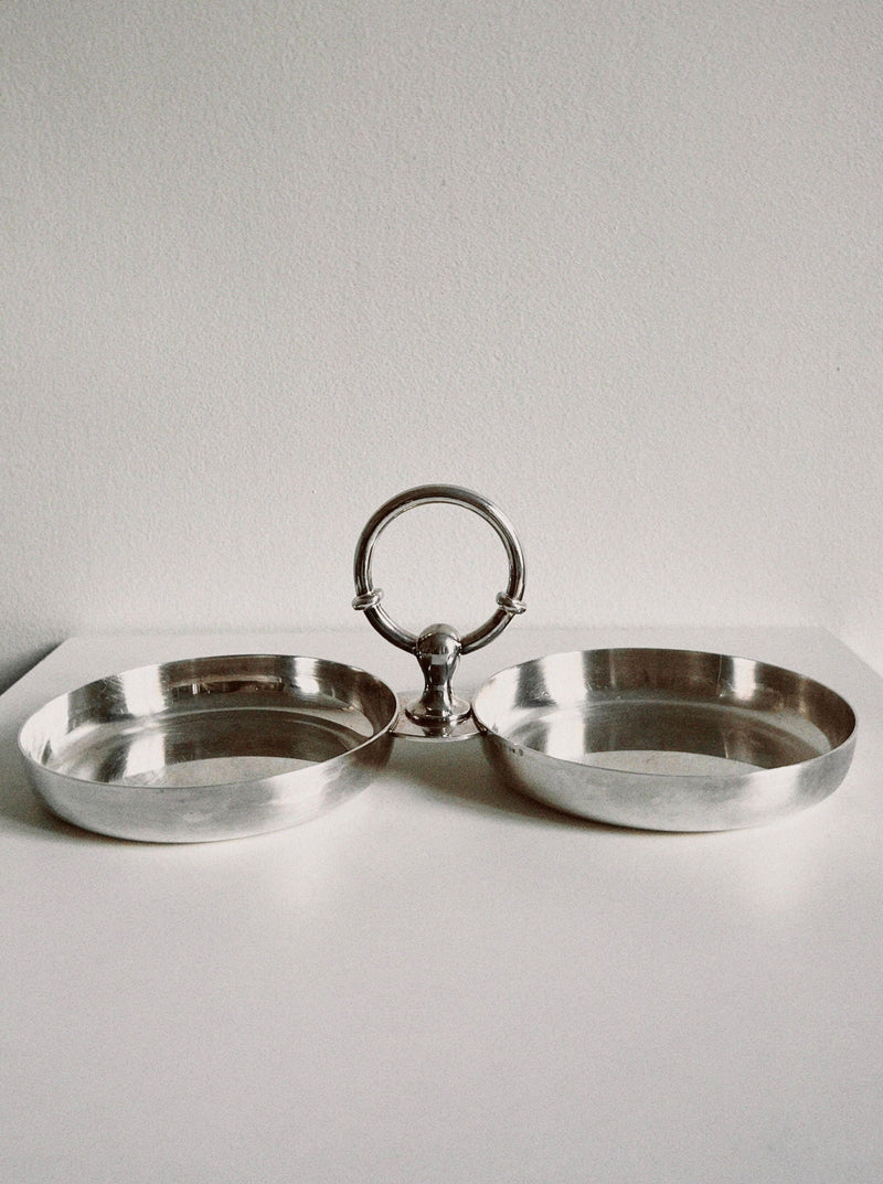 The Manushi Double Silver Server, featuring a central ring handle, is displayed elegantly on a white surface against a plain, light background.
