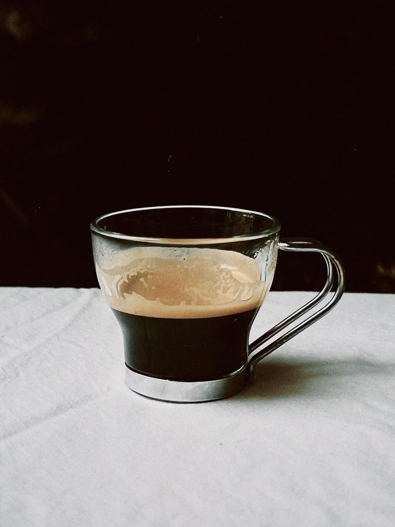 The Manushi Set of 6 French Espresso Cups features glass mugs with metallic handles, filled with dark coffee and crema, displayed on white fabric against a dark background.