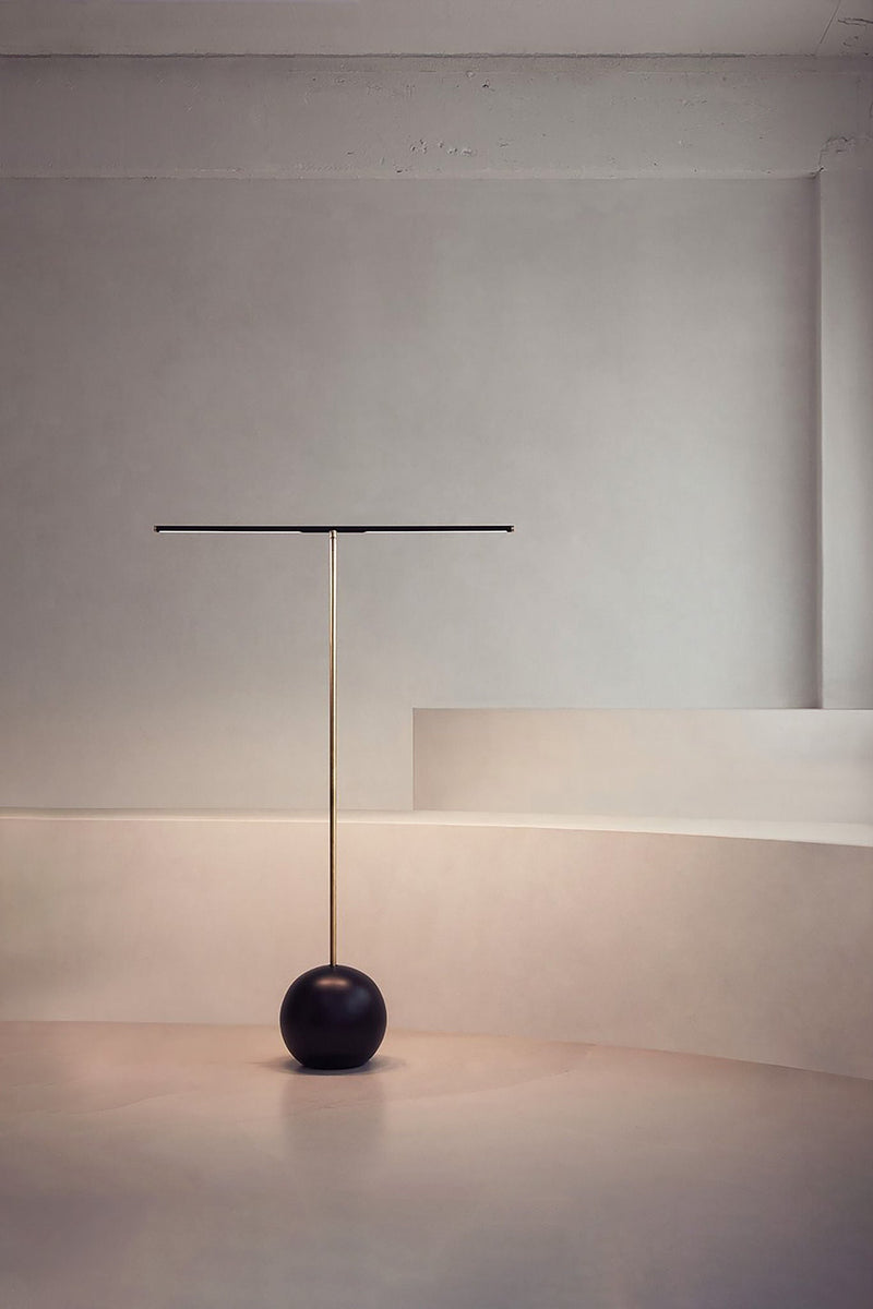 The 'T-Lamp' Floor Lamp by Heako Studio features a thin horizontal light bar atop a tall brass pole with a black spherical base, adding sculptural illumination to a softly lit, neutral-toned modern room.