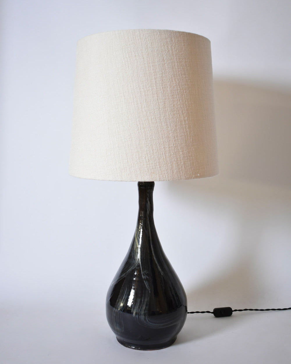 The Black Ivory Brush Lamp, Helder by Barracuda Interiors features a glossy dark ceramic teardrop base, a large textured off-white shade, and a black power cord for a modern touch.