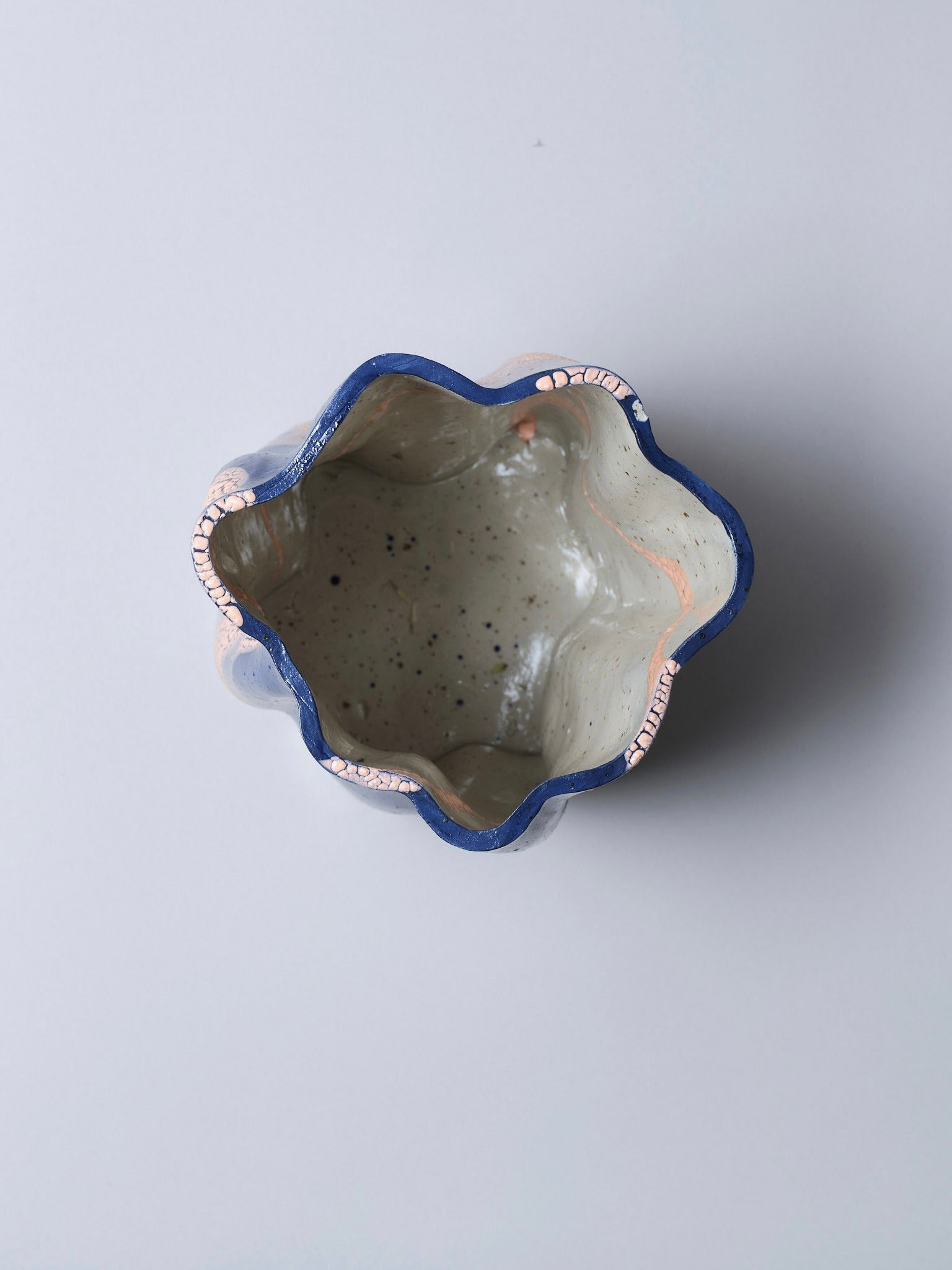 Top view of Astrid Öhman’s Swirly Vase Blue and Peach: a handmade stoneware vase with a wavy, dark blue rim accented by pink cracks and a light speckled glaze lining the interior, set against a plain background.