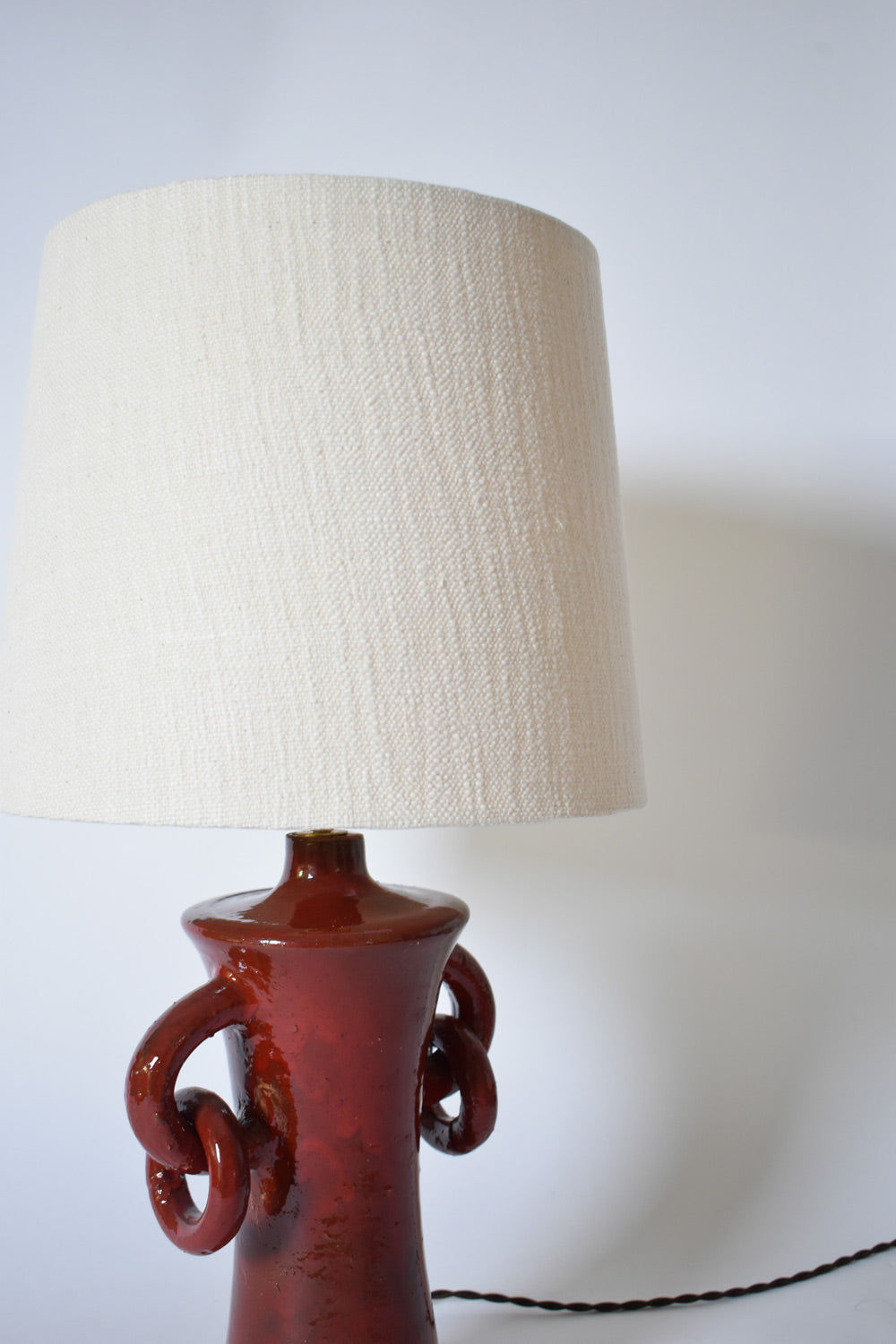 The "Kasaï" Red Handles Ceramic Table Lamp by Barracuda Interiors features a glossy reddish-brown ceramic base with decorative handles and a textured off-white fabric shade, shown against a plain white background.
