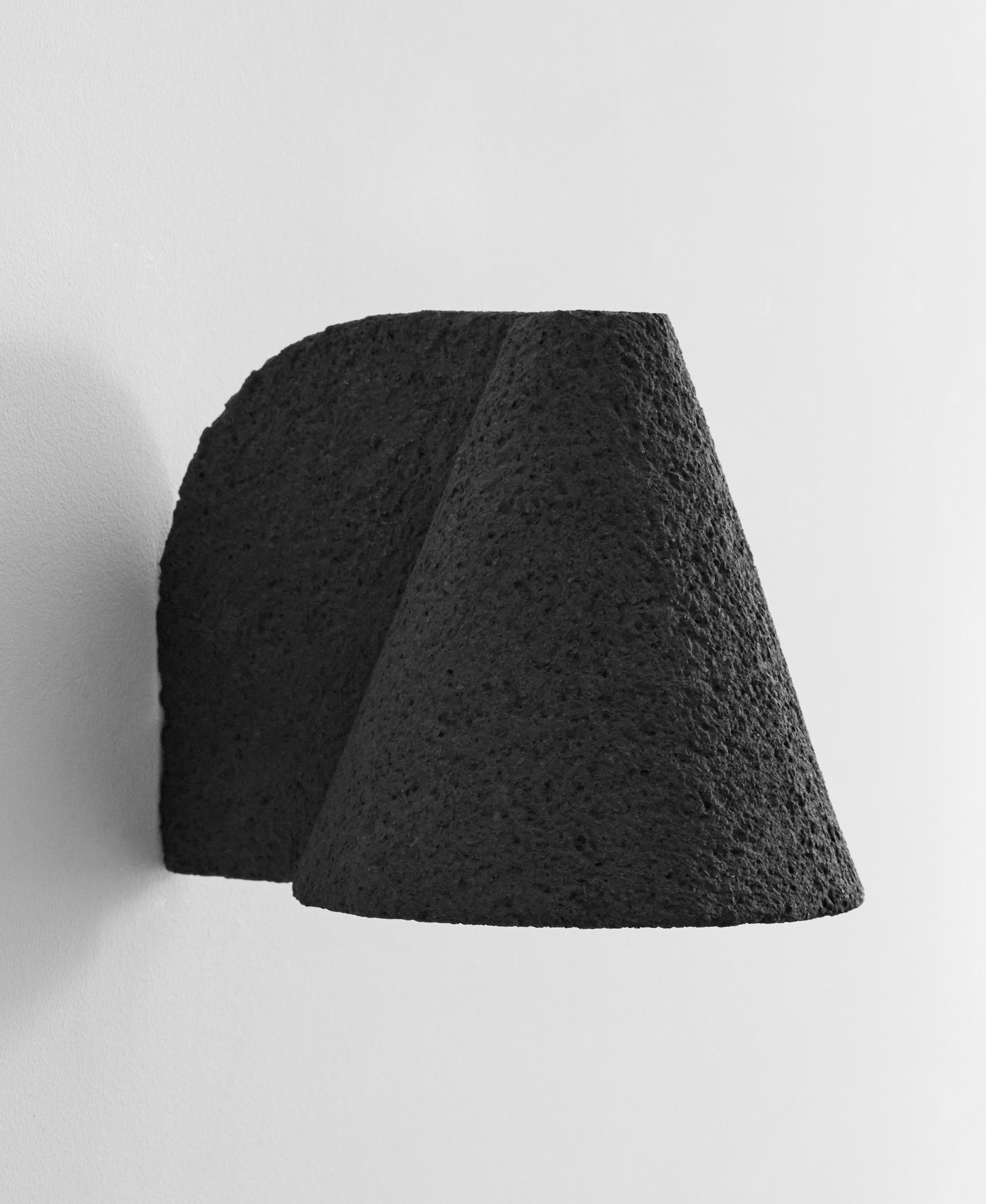 The Esoteric Granular Wall Lamp by Mariza Galani, featuring a black textured cone shape and rough finish, is mounted on a white wall and casts a soft shadow to the left.