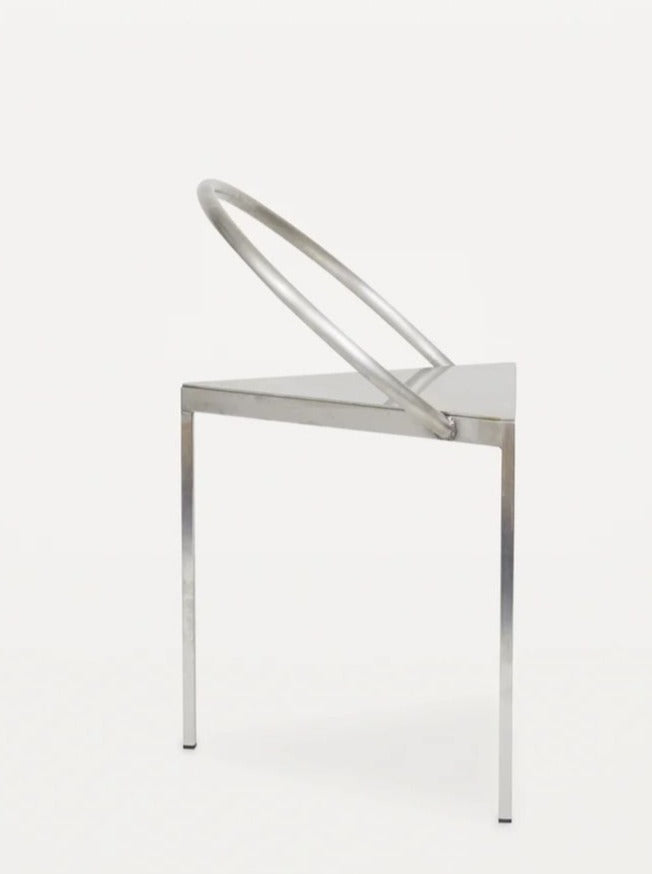 The FRAMA Triangolo Chair | Stainless Steel features a minimalist triangular seat, three straight legs, and an angled circular backrest—a striking modern piece set against a white background.