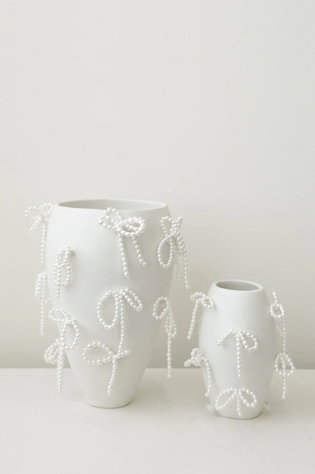 Two "Notsobig" Power, Corruption, Lies Small Vases in matte white by Completedworks, each adorned with faux pearl bows, are displayed side by side against a plain background—striking sculptural pieces for contemporary decor.