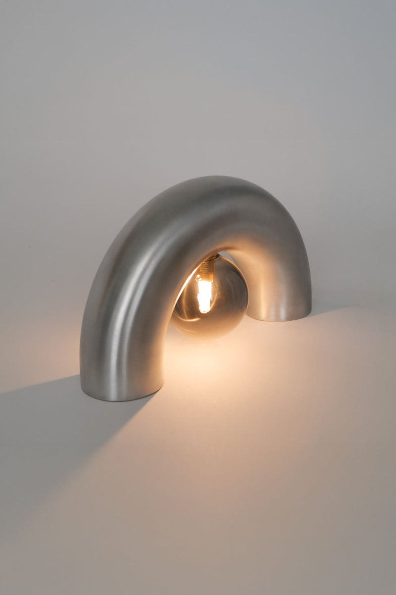 The Arcora Underglow Aluminum Arc Lamp by Heako Studio features a modern, upside-down U design that arches over a glowing bulb, casting a soft, warm light on any surface to create a calming atmosphere.