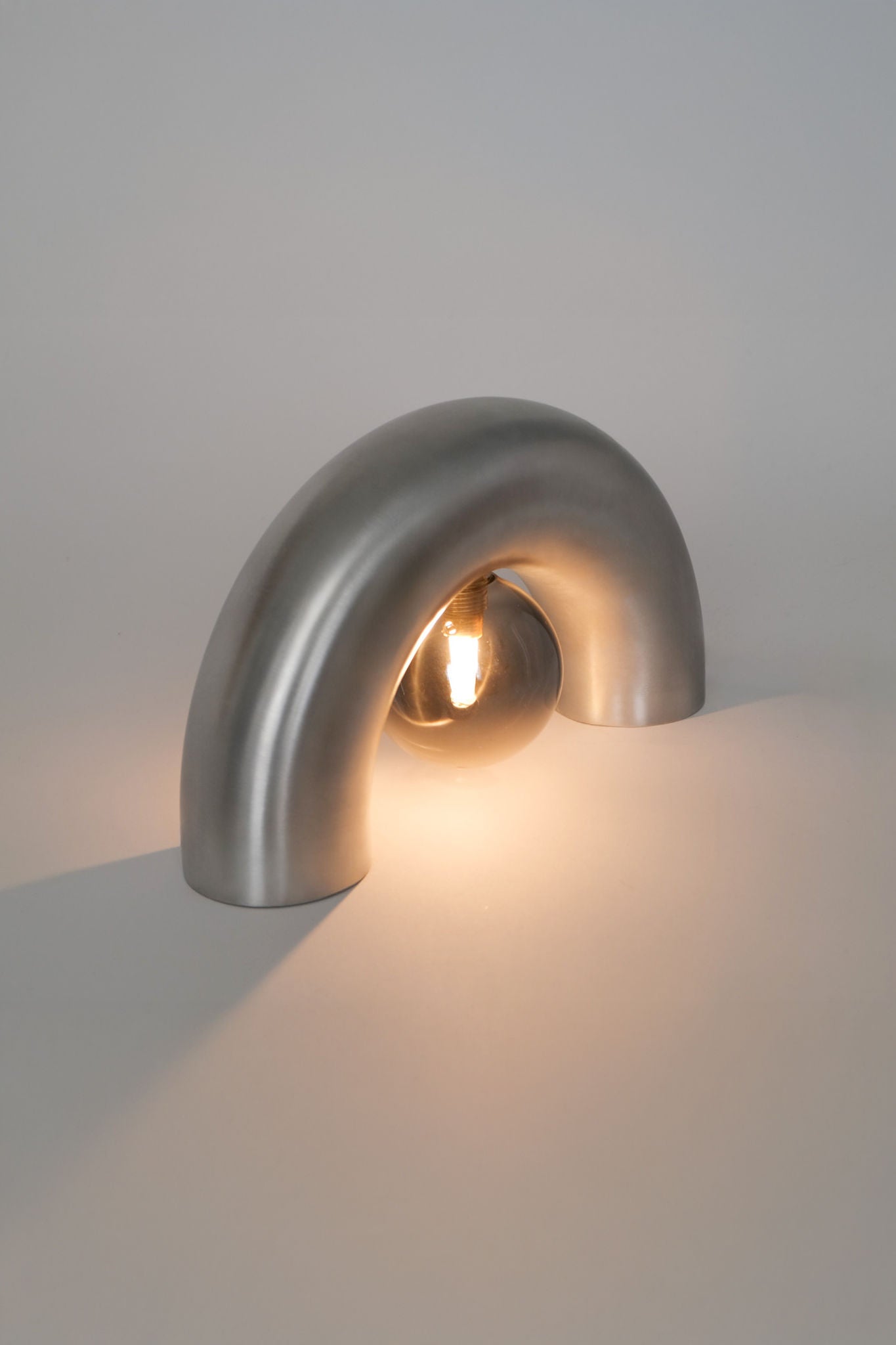The Arcora Underglow Aluminum Arc Lamp by Heako Studio features a modern, upside-down U design that arches over a glowing bulb, casting a soft, warm light on any surface to create a calming atmosphere.