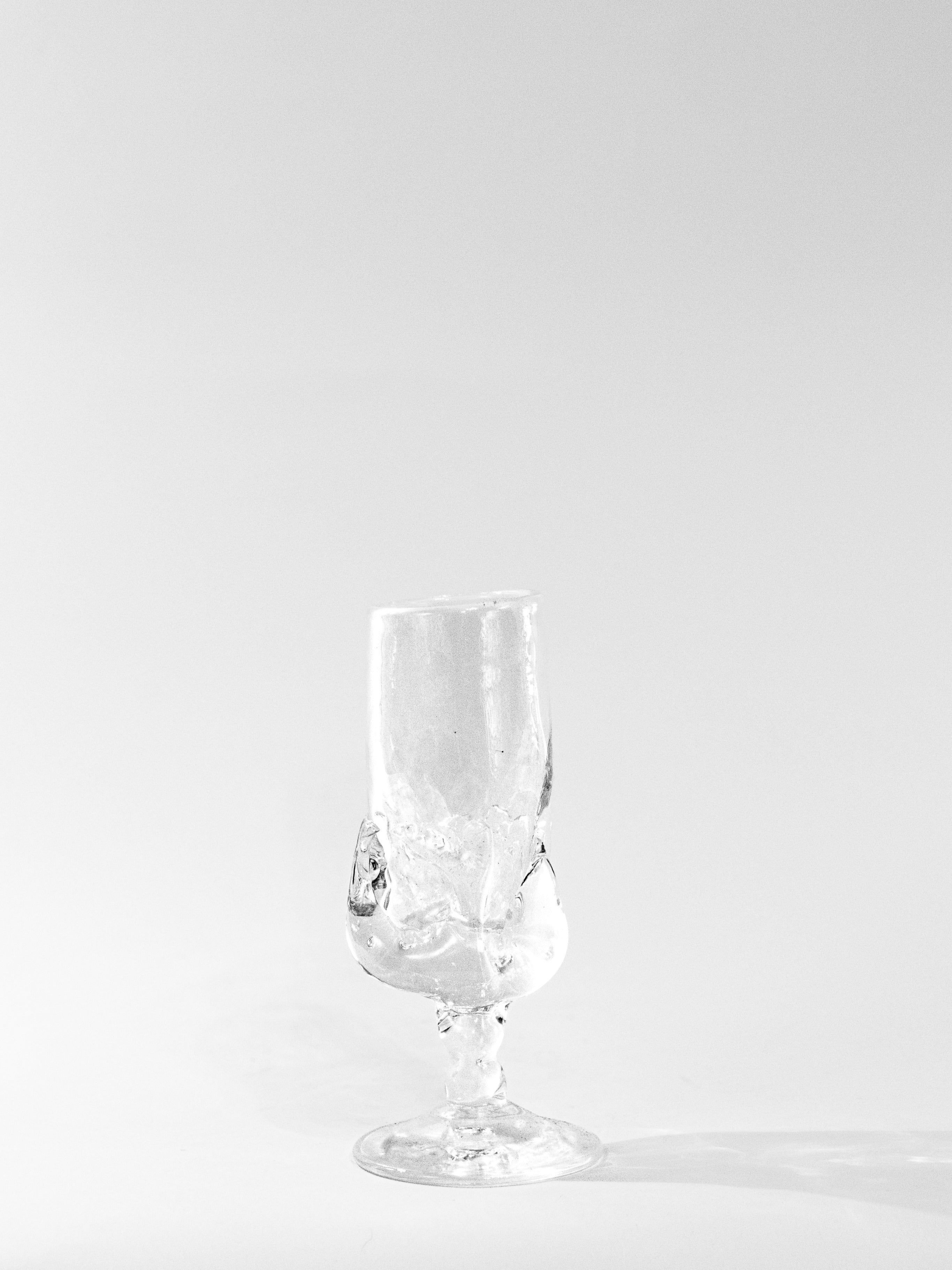 A clear, irregularly shaped Table Goblet by Szkło Studio stands on a plain white background, casting a rightward shadow. Its twisted, abstract form showcases expert glass blowing and appears empty.