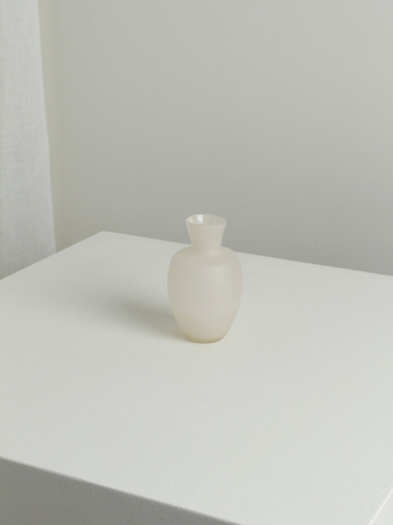 The Small Vase in Moon by Verre d'Onge, featuring a frosted white round body and narrow neck, is centered on a white surface with a light gray background in a minimalistic, softly lit setting.