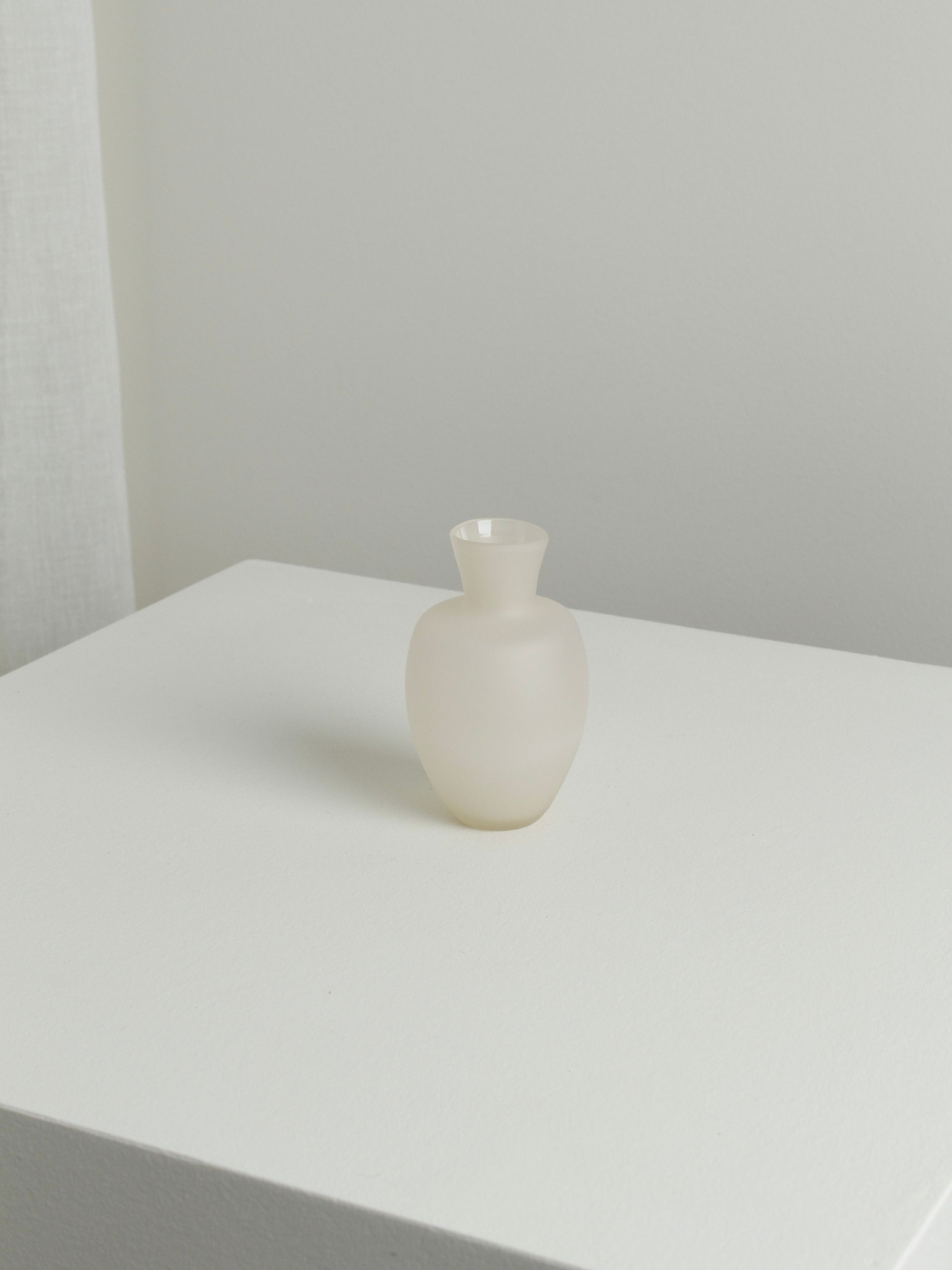 The Small Vase in Moon by Verre d'Onge, featuring a frosted white round body and narrow neck, is centered on a white surface with a light gray background in a minimalistic, softly lit setting.