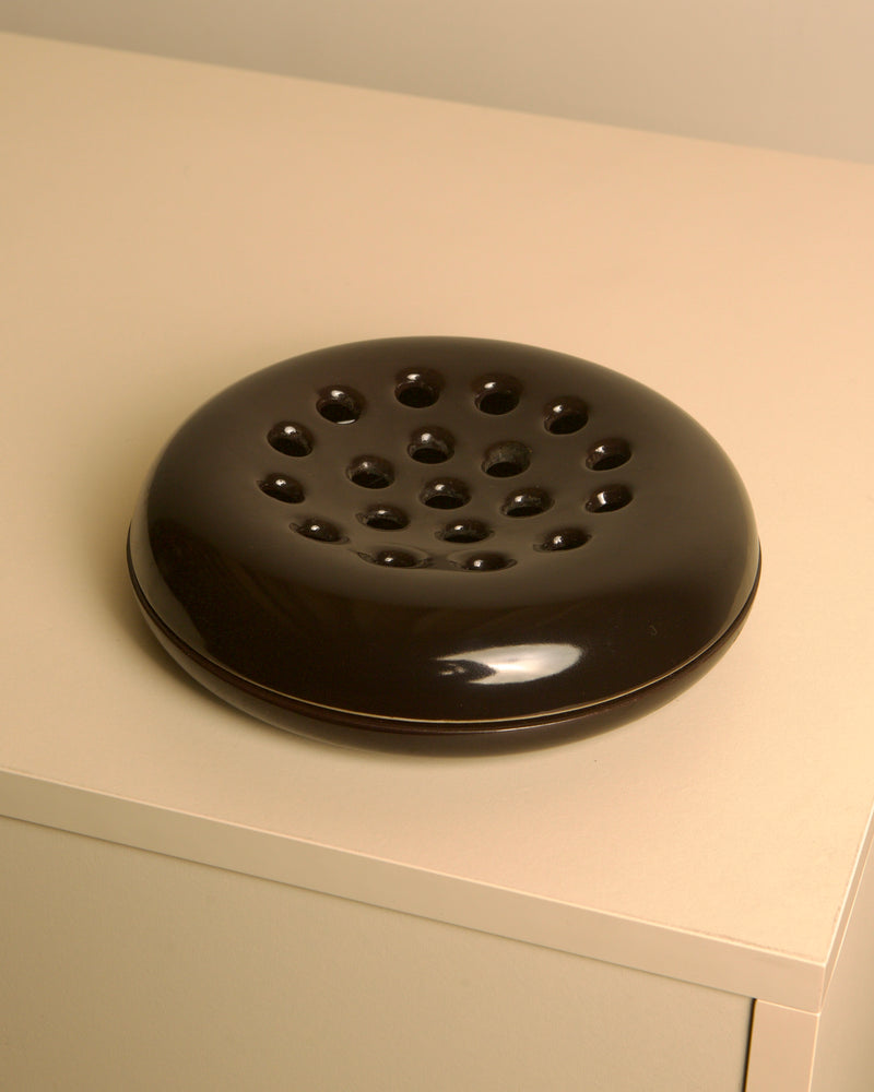 A Treaptyque Italian Brown Ceramic Ashtray by Angelo Mangiarotti for Brambilla, 1960s, featuring a round, glossy finish and multiple top holes, rests on a light beige surface.