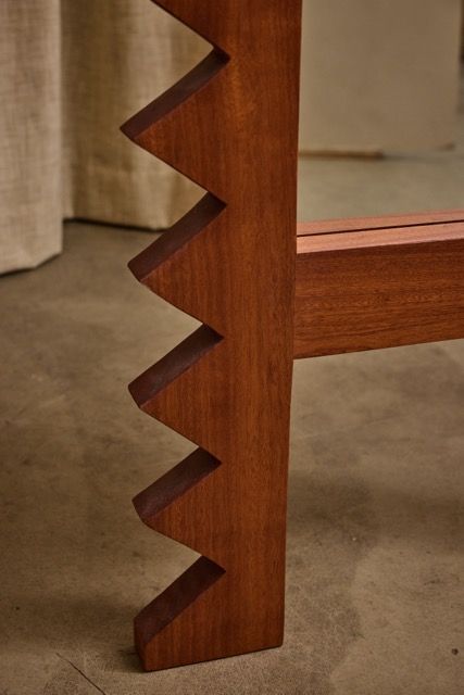 Close-up of the Jura Mirror by Ah Um Design Studio, showcasing its solid mahogany frame with a zigzag edge detail, set against a concrete floor and neutral fabric backdrop.